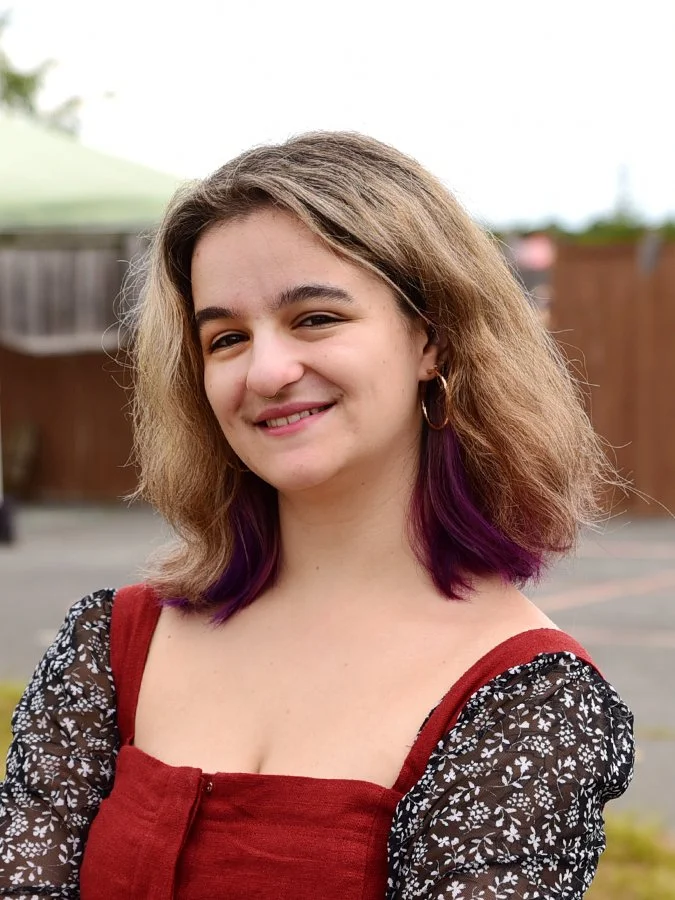 Young woman with shoulder-length wavy hair, brown with purple tips, smiling outdoors in front of a wooden fence and green canopy.