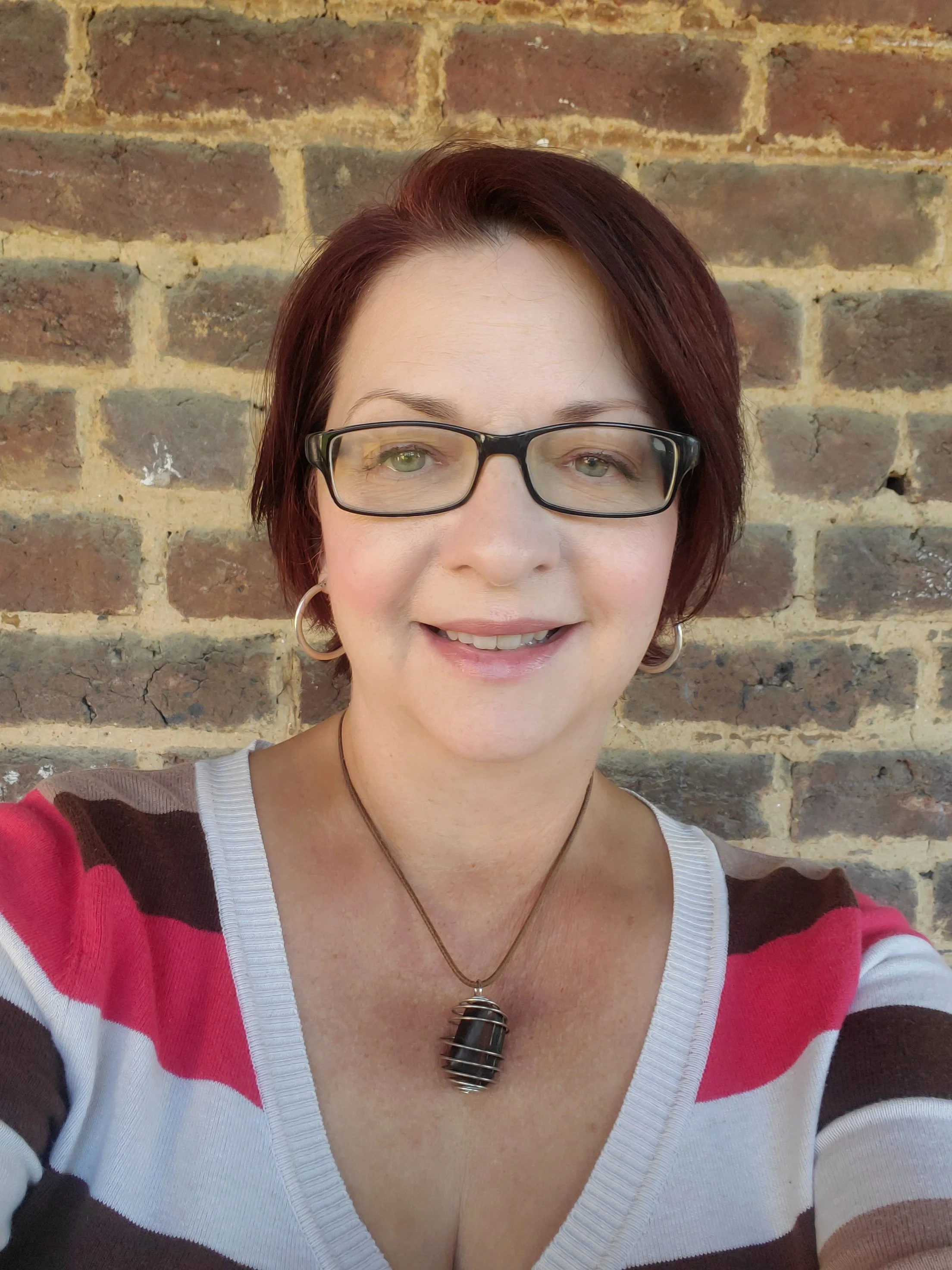 Close-up of a woman with short reddish-brown hair, wearing glasses, a striped sweater in red, white, and brown, a pendant necklace, and earrings, standing in front of a brick wall.