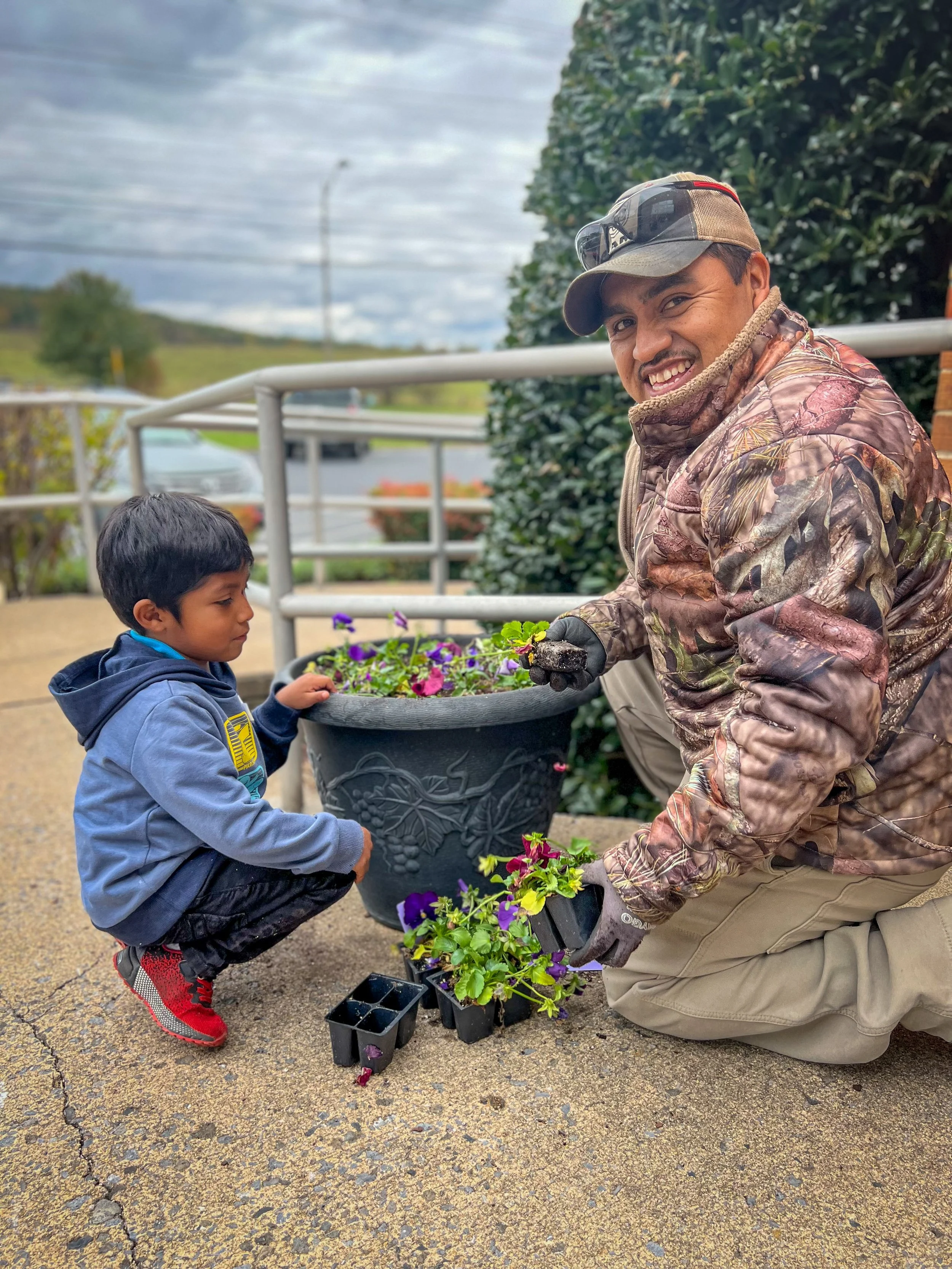 A man and a young boy planting purple and yellow flowers in a large black pot outdoors on a cloudy day. The man is wearing camouflage clothing, a cap, and sunglasses, smiling at the camera. The boy is wearing a blue jacket and red shoes, focused on planting flowers.