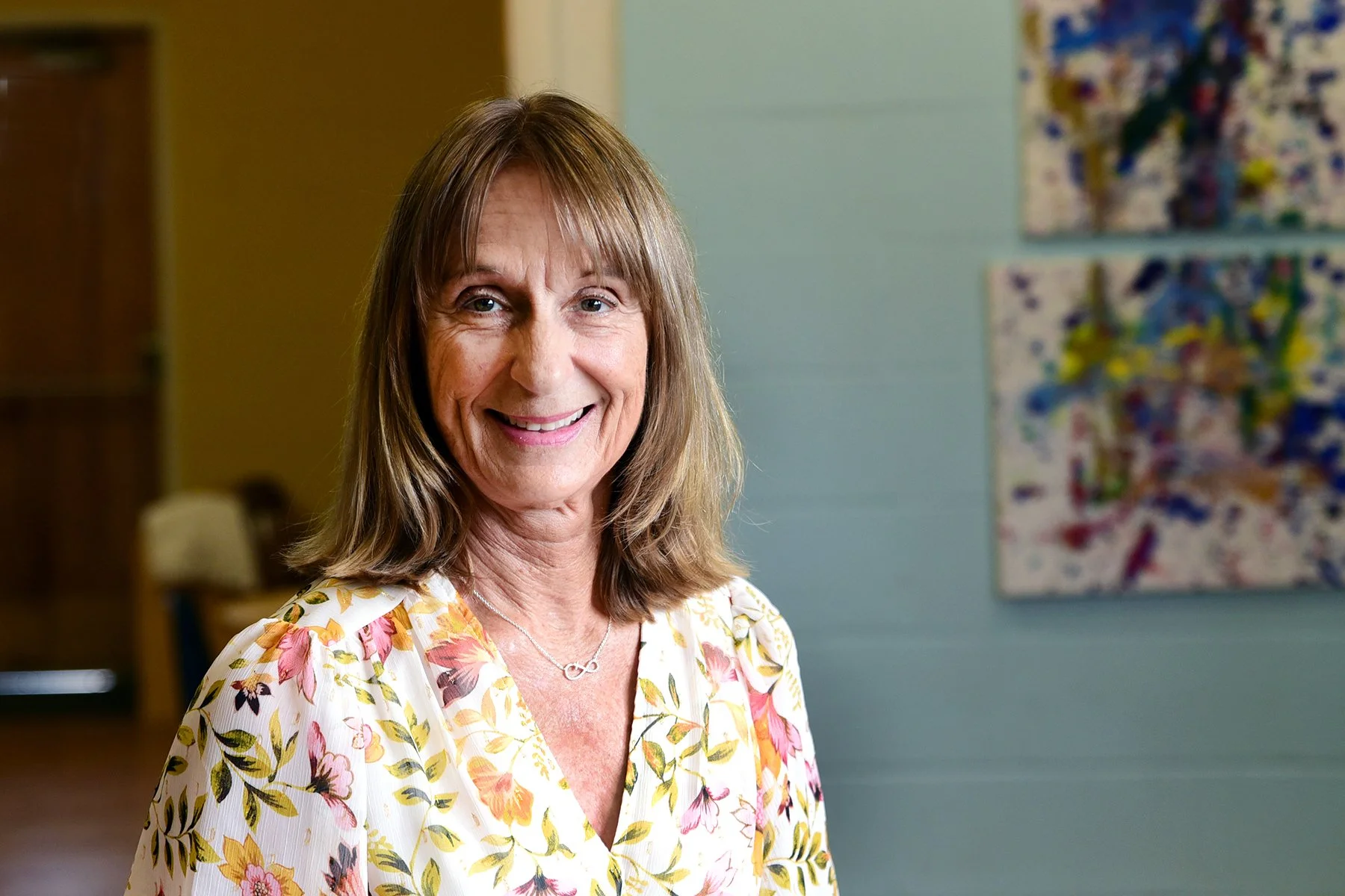 Smiling woman with shoulder-length brown hair wearing a floral blouse and a silver infinity necklace, standing indoors in front of a colorful abstract painting on a light blue wall.