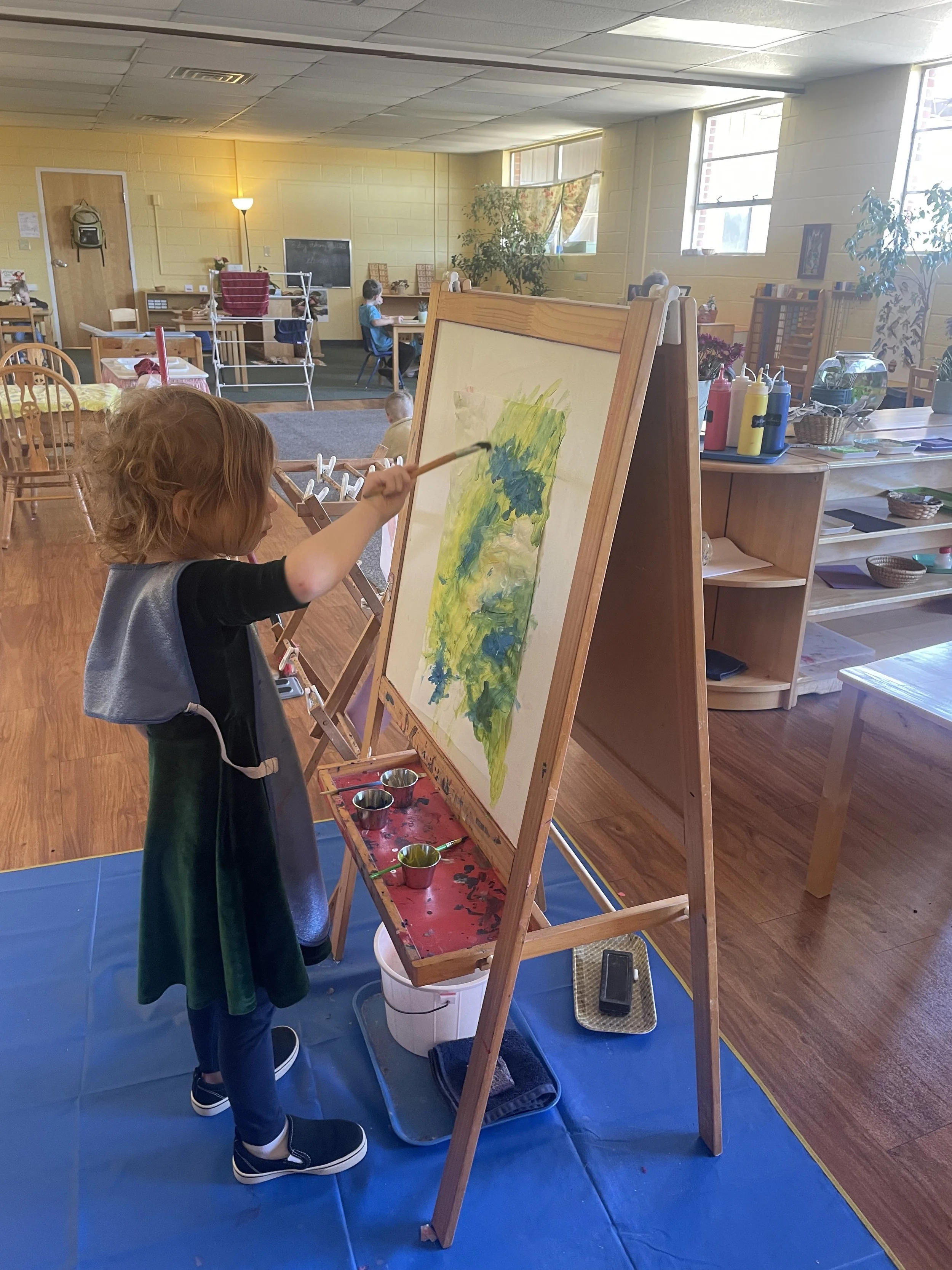 A young child painting on a canvas with green and yellow colors in an art classroom. The child is standing and wearing a blue apron. The classroom has wooden furniture, plants, and windows letting in natural light.