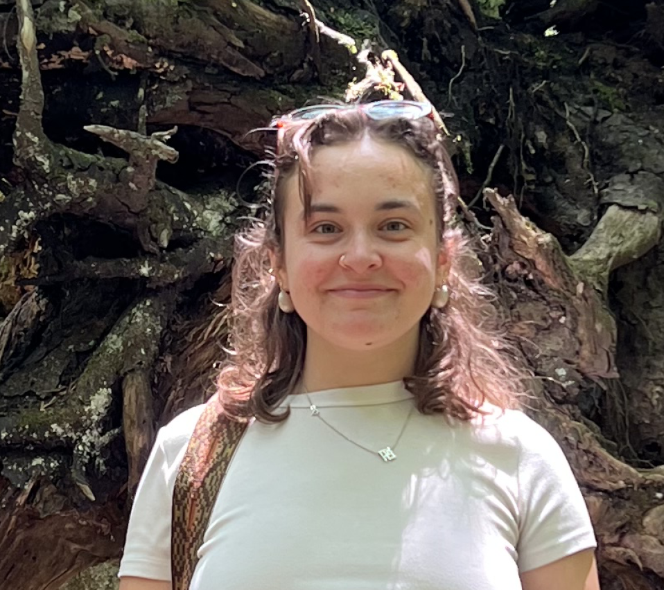 A young woman with curly hair and a nose ring smiling outdoors in front of tree roots, wearing a white t-shirt, sunglasses on her head, pearl earrings, and a necklace.