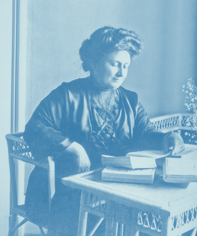Maria Montessori reading a book at a wicker table.