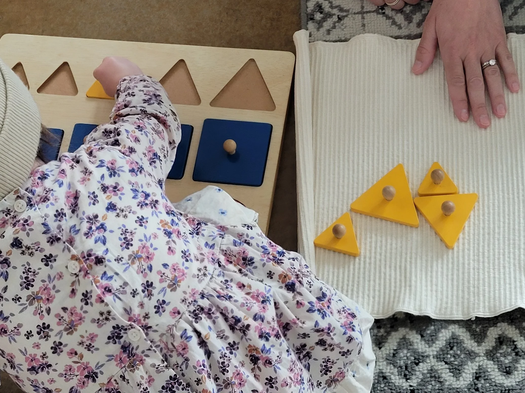 A child wearing a floral dress is reaching for yellow wood geometric shapes on a wooden table next to an adult's hand resting on a yellow cloth with more yellow wood shapes. The table is set on a rug and a carpet.