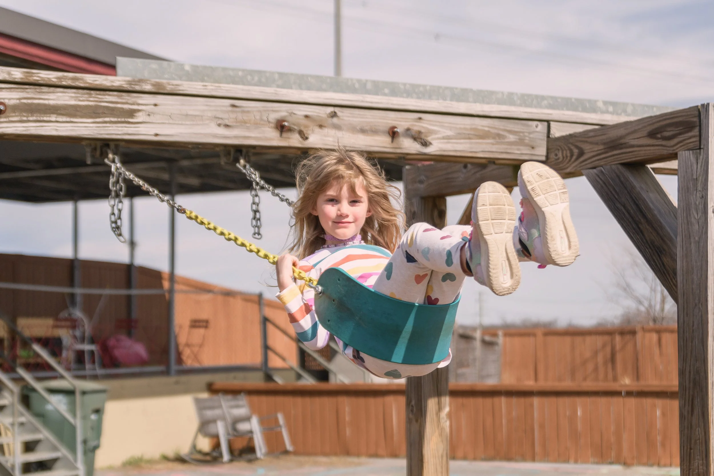 Young girl with blonde hair playing on a swing set outdoors, wearing colorful clothing with hearts, in a backyard or playground area with a wooden fence and storage shed in the background.
