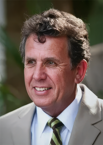 A middle-aged man with curly brown hair, wearing a beige suit, white shirt, and green striped tie, smiling outdoors.