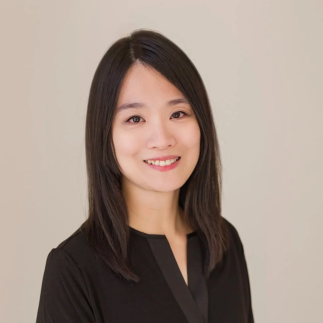 A woman with shoulder-length dark hair smiling at the camera, wearing a black top against a plain beige background.