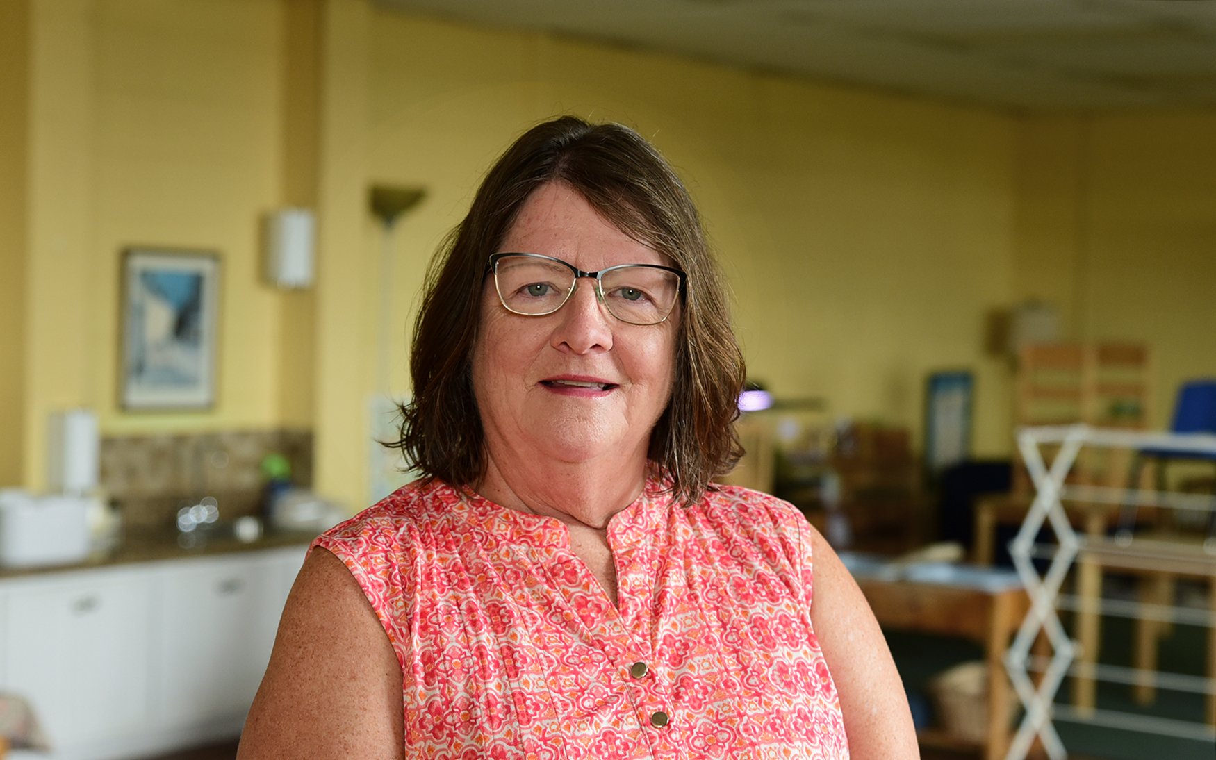 A woman with glasses wearing a pink patterned sleeveless blouse, standing in a room with yellow walls and various furniture and decorations in the background.
