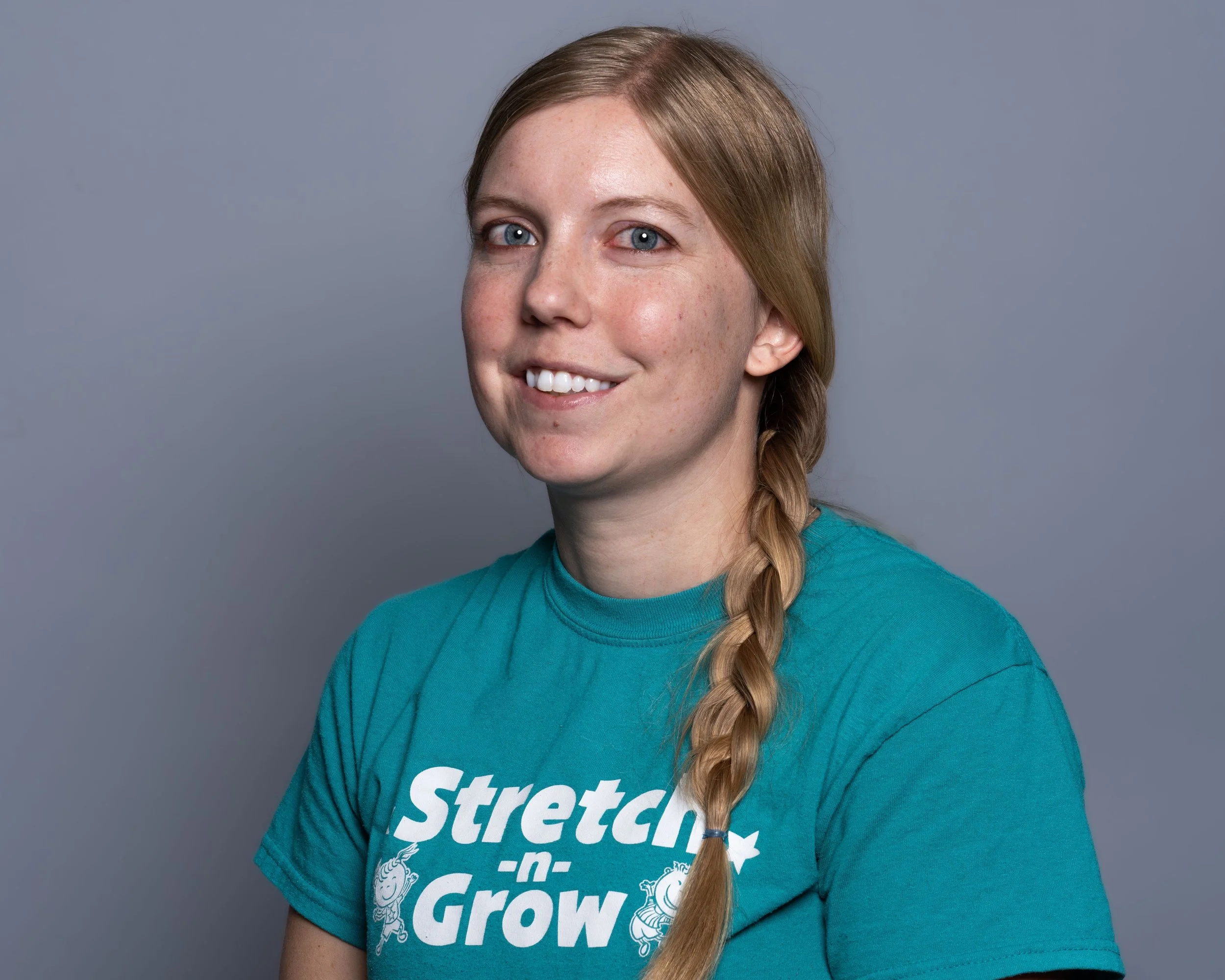 A young woman with long blond hair styled in a braid, smiling, wearing a teal T-shirt with white text and graphics, sitting against a plain grey background.