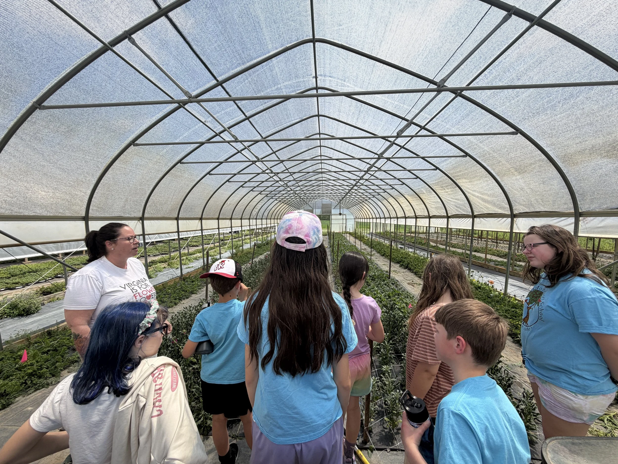 Group of children and adults inside a large greenhouse with plants growing in rows, under a curved, transparent roof.