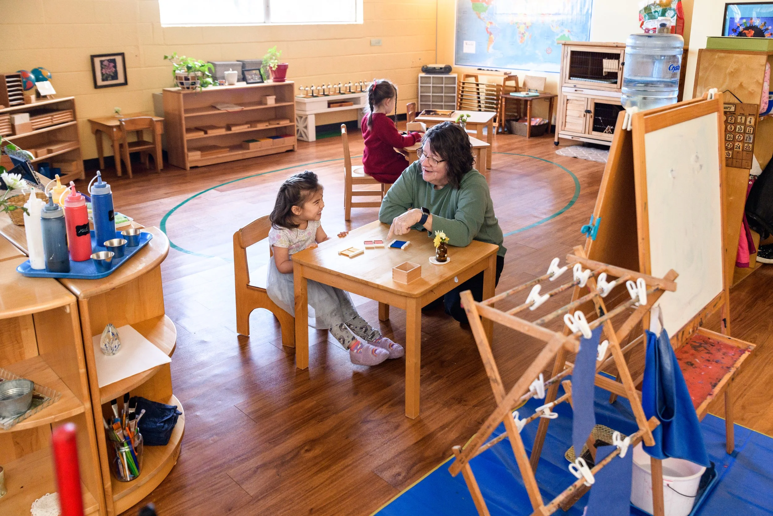 A classroom scene with children and a teacher. A young girl is sitting at a small table with a teacher, engaging in a conversation. A girl in a red dress is sitting at another table, focusing on an activity. The room has wooden furniture, educational materials, and a world map on the wall.