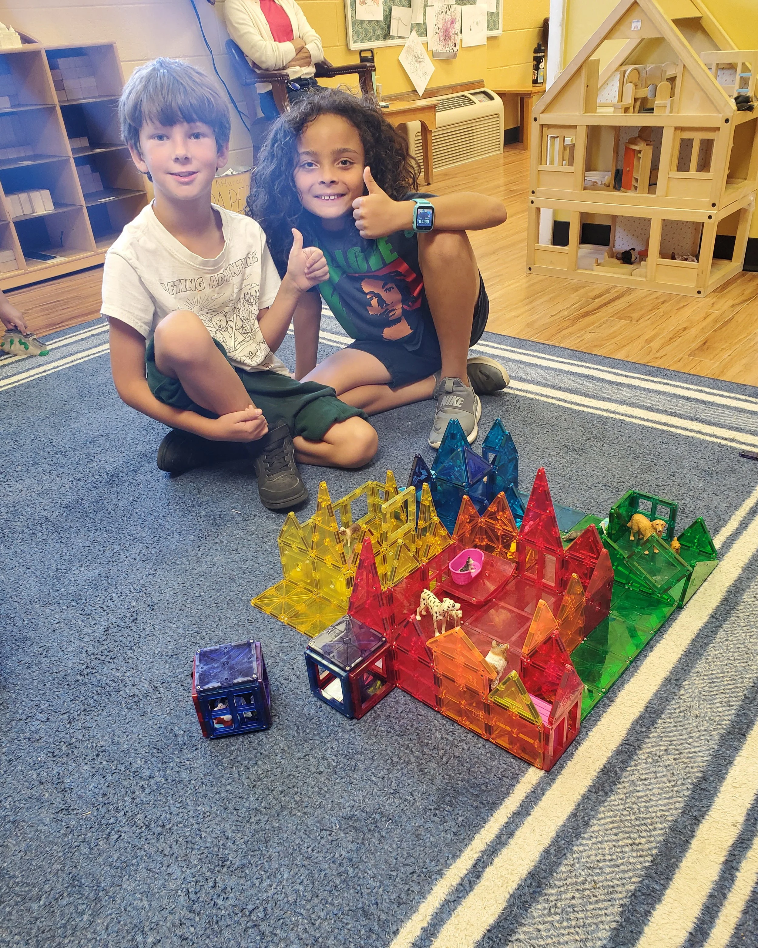 Two children, a boy and a girl, sitting on a blue carpet in a classroom, surrounded by colorful magnetic building blocks arranged in the shape of a house.
