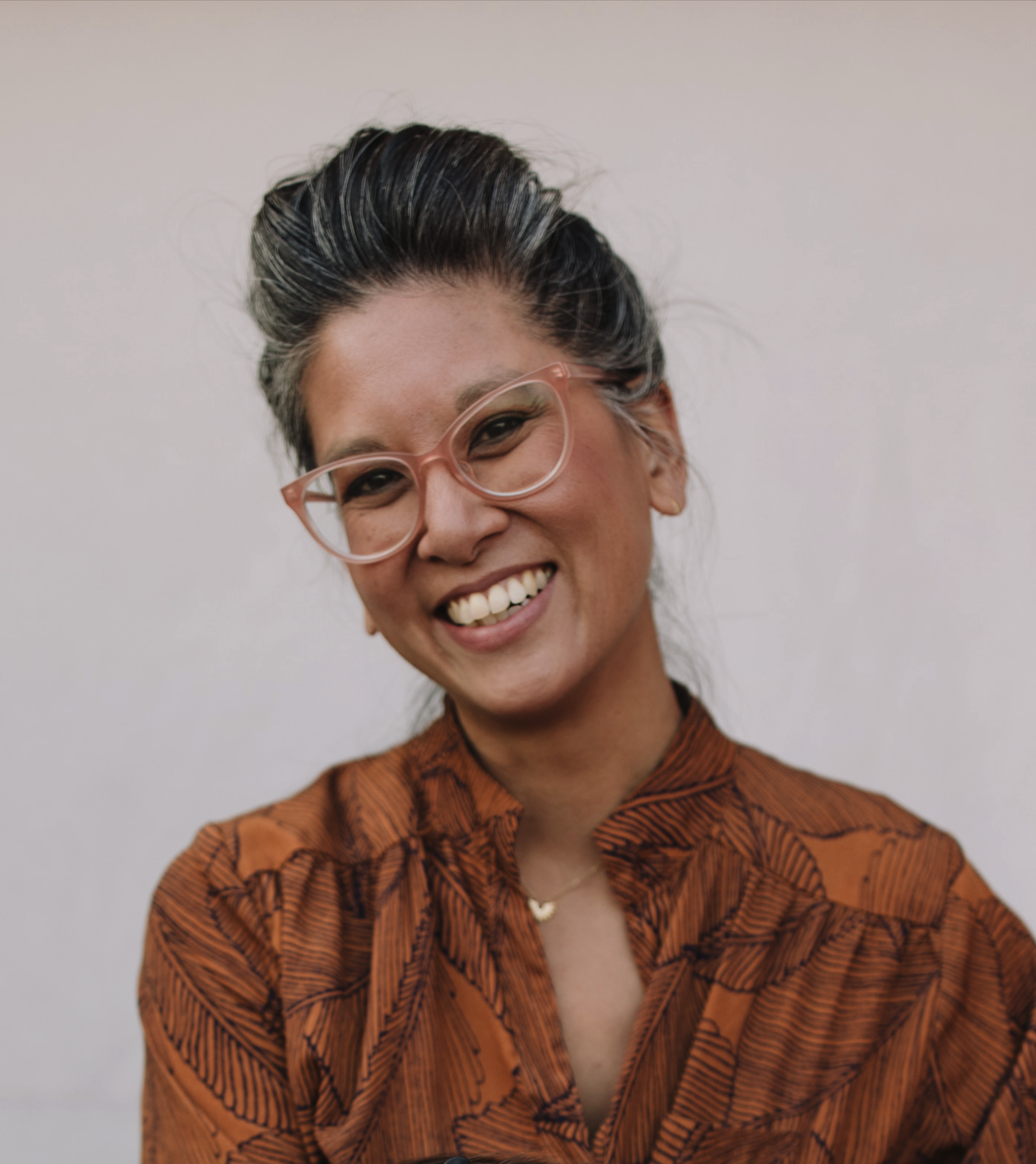 A smiling woman with glasses and gray and black hair, dressed in a brown patterned shirt.