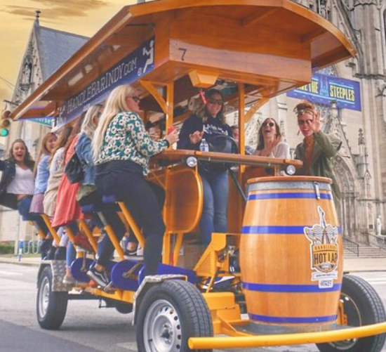A group of people riding in a pedal-powered beer barrel trolley on a city street.