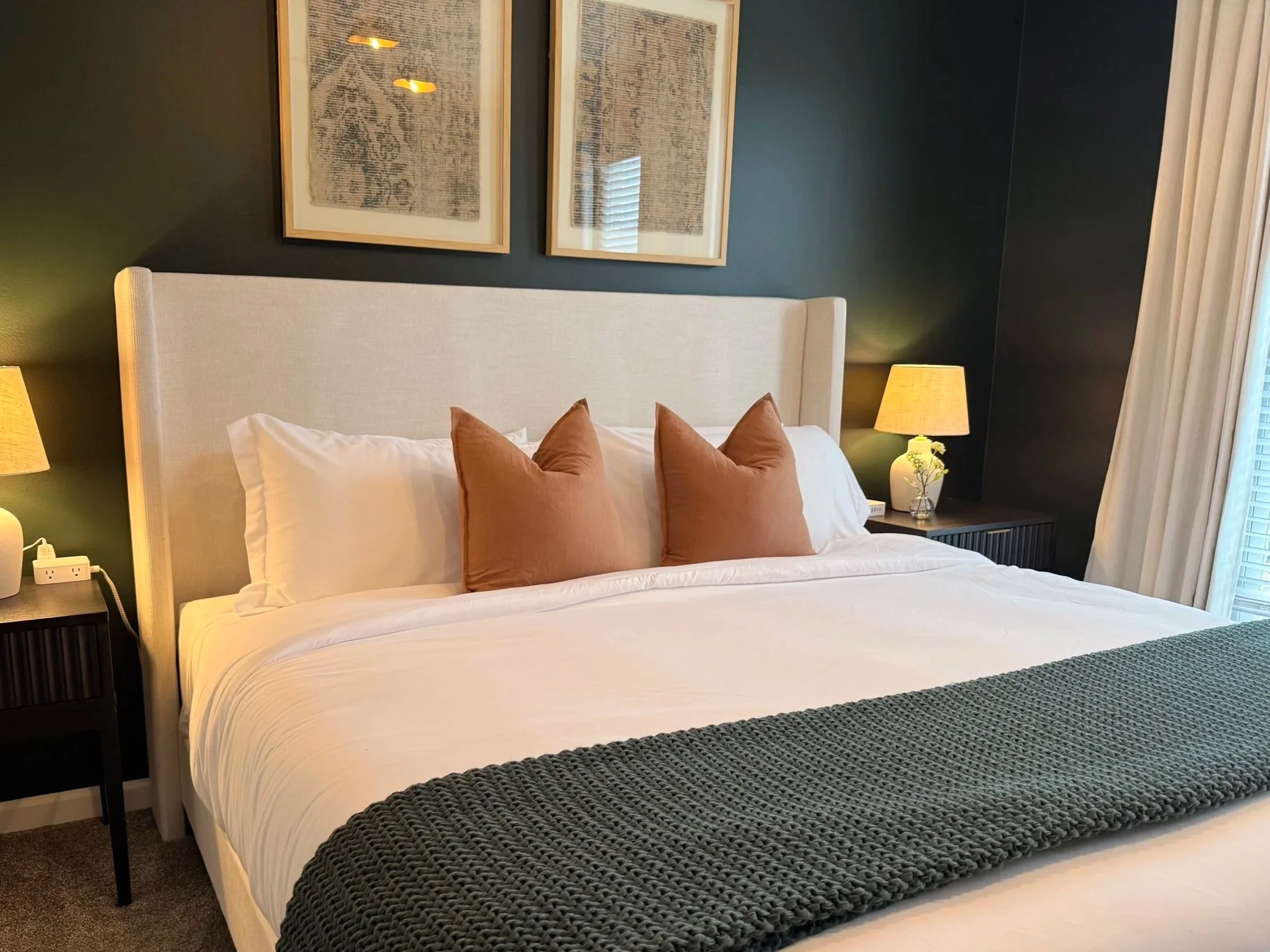 A neatly made bed with white linens, two white pillows, and two brown decorative pillows, flanked by two bedside tables with matching lamps, in a modern bedroom with dark painted walls and framed artwork above the bed.