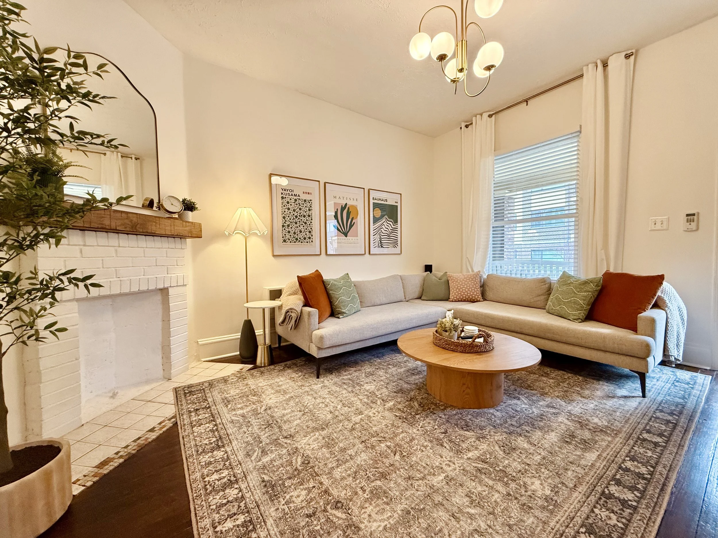 Living room with a white sofa, green and orange throw pillows, a round wooden coffee table, a white brick fireplace with a wooden mantle, framed artwork on the wall, a tall floor lamp, large windows with white curtains, and a decorative area rug.