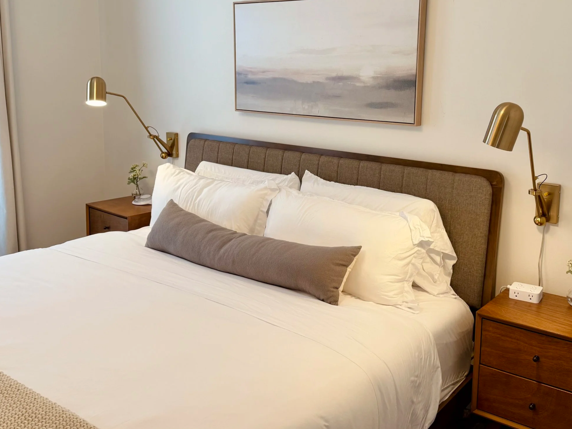 A neatly made bed with white linens, multiple white pillows, a long gray bolster pillow, a wooden headboard, and two matching gold wall-mounted reading lamps. There are wooden nightstands on each side with a small plant on the left and an outlet with a plug on the right. An abstract painting hangs above the bed.