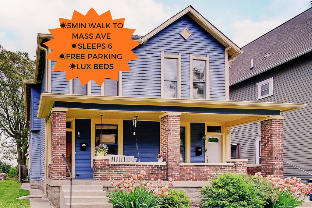 A blue two-story house with a front porch supported by red brick columns, surrounded by green plants and pink flowers. An orange starburst-shaped sign highlights features like being a five-minute walk to Mass Ave, sleeping six, free parking, and luxury beds.