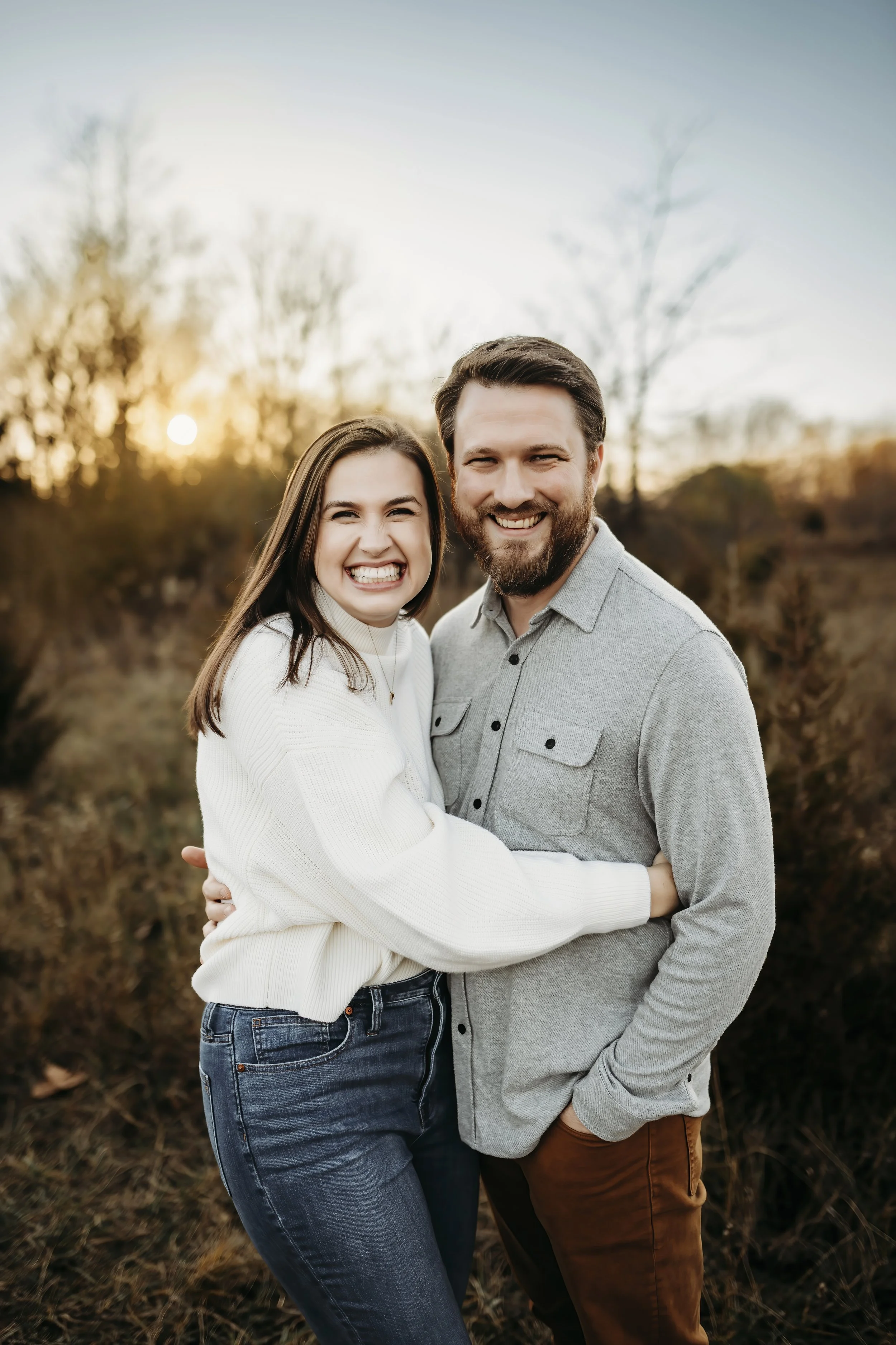 A happy couple embracing outdoors during sunset, smiling at the camera with trees and a setting sun in the background.
