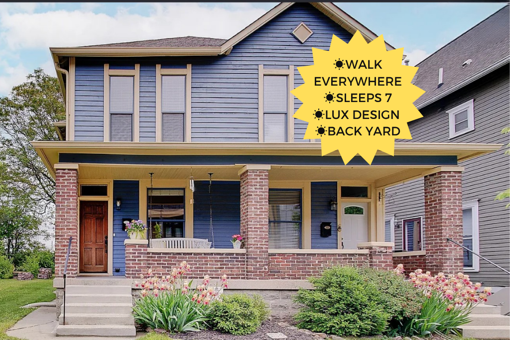 Front view of a two-story blue house with a covered porch, brick columns, and a small lawn with flowers. A yellow starburst text overlay lists features: walk everywhere, sleeps 7, luxury design, backyard.