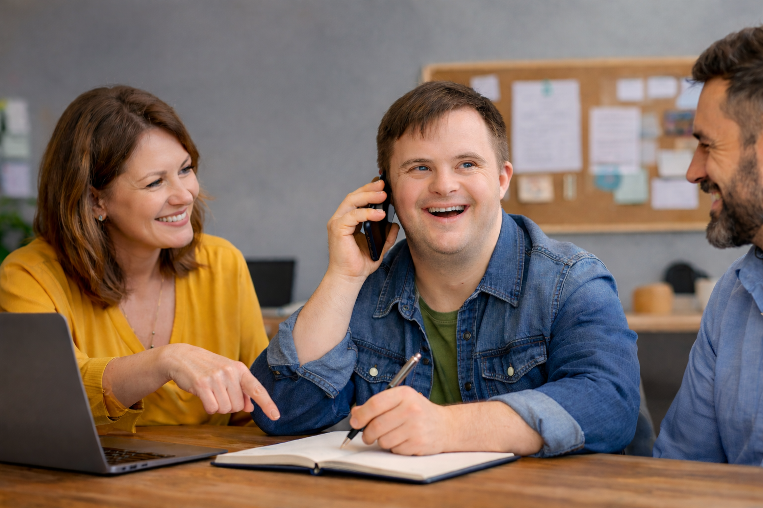 A man talking on the phone and writing in a notebook while sitting with two smiling colleagues at a table.