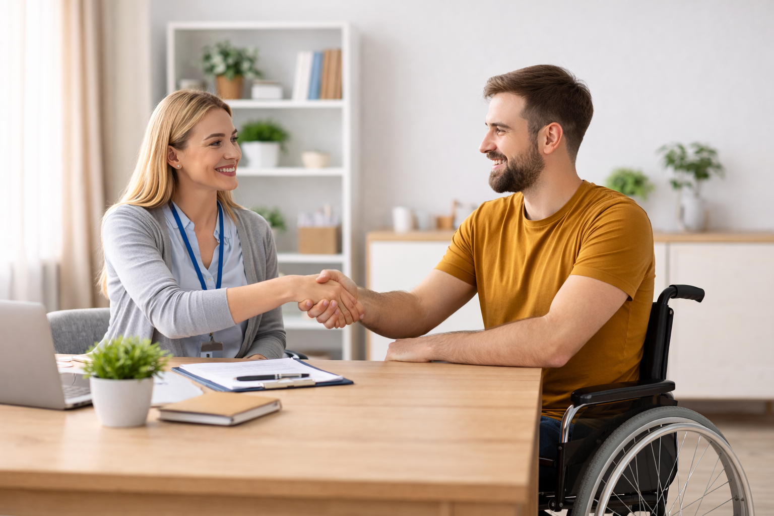 A support worker and a man using a wheelchair smiling and shaking hands across a desk.