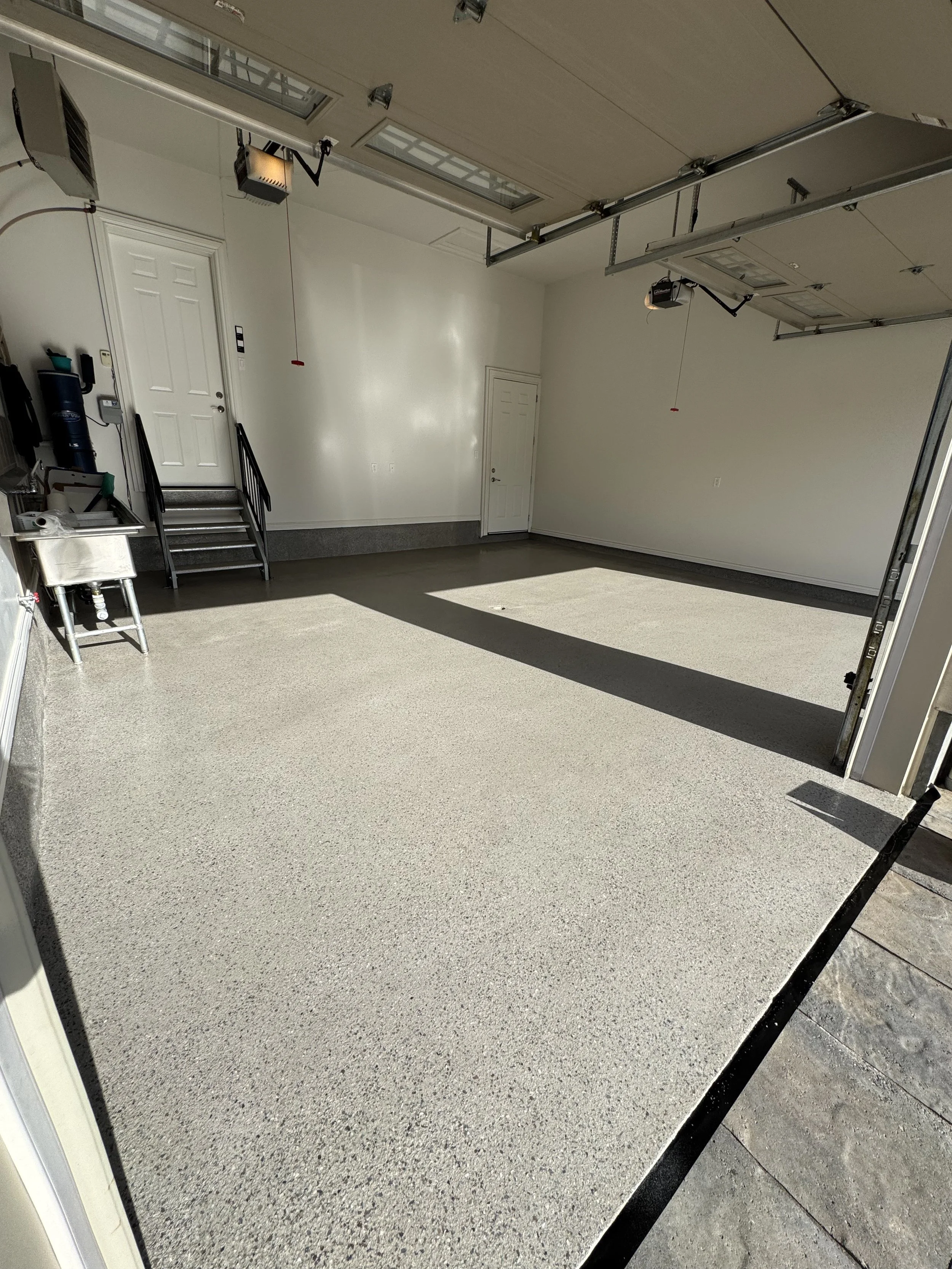 Empty garage with light-colored speckled epoxy floor, white walls, a small sink on the left, and two garage door openers on the ceiling.