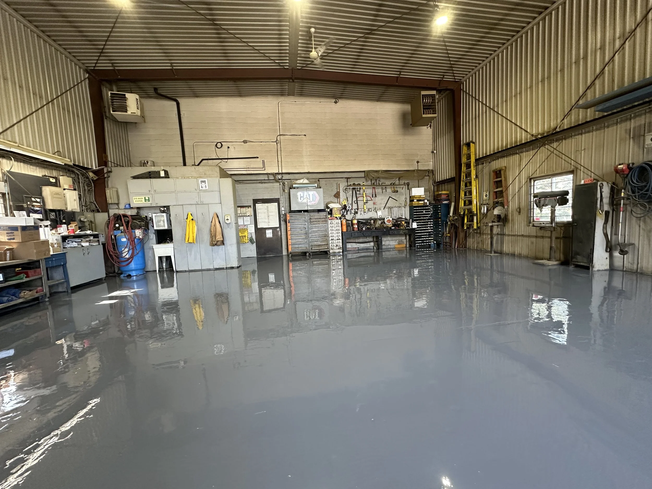 Interior of a workshop or garage with a freshly painted, shiny gray or silver concrete floor, various tools, equipment, and storage cabinets along the walls, a ladder, and windows letting in natural light.