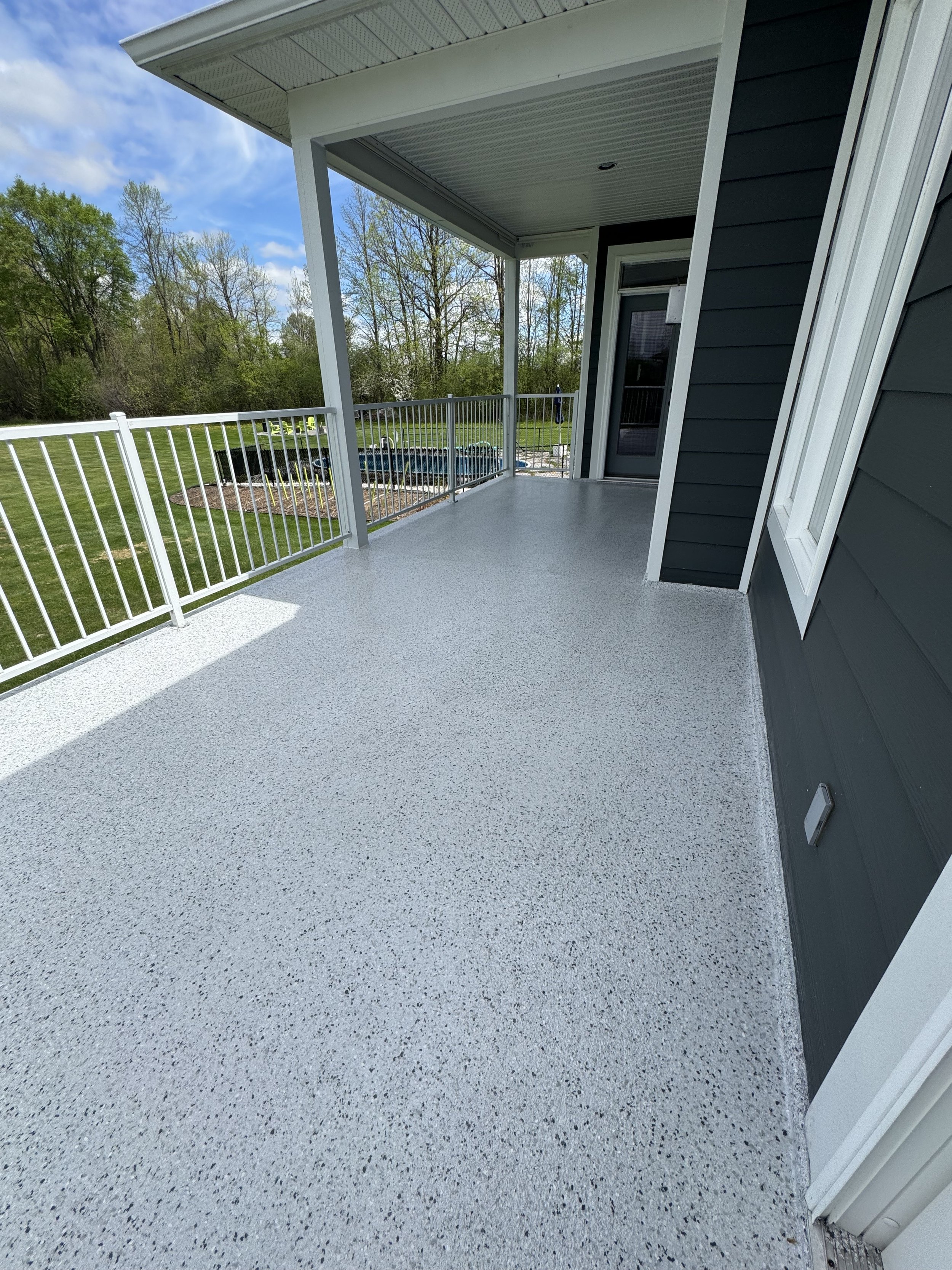 Empty outdoor balcony with gray speckled flooring, white railing, and a dark gray house wall with a window and door, overlooking a backyard with trees and an above ground pool.