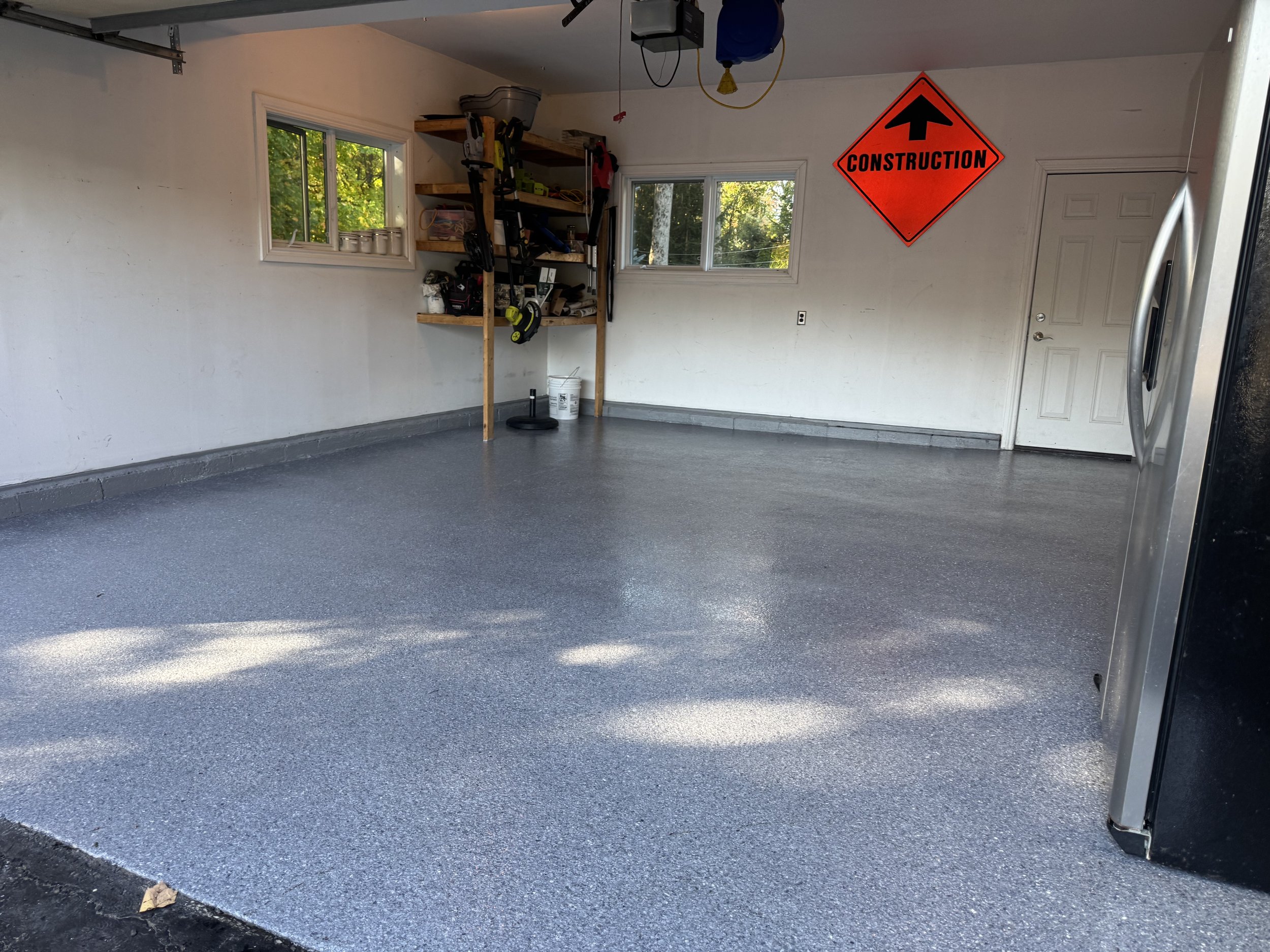 Empty garage with newly painted or epoxy coated floor, white walls, two small windows, shelving unit with tools, and a bright orange 'Construction' sign on the wall.