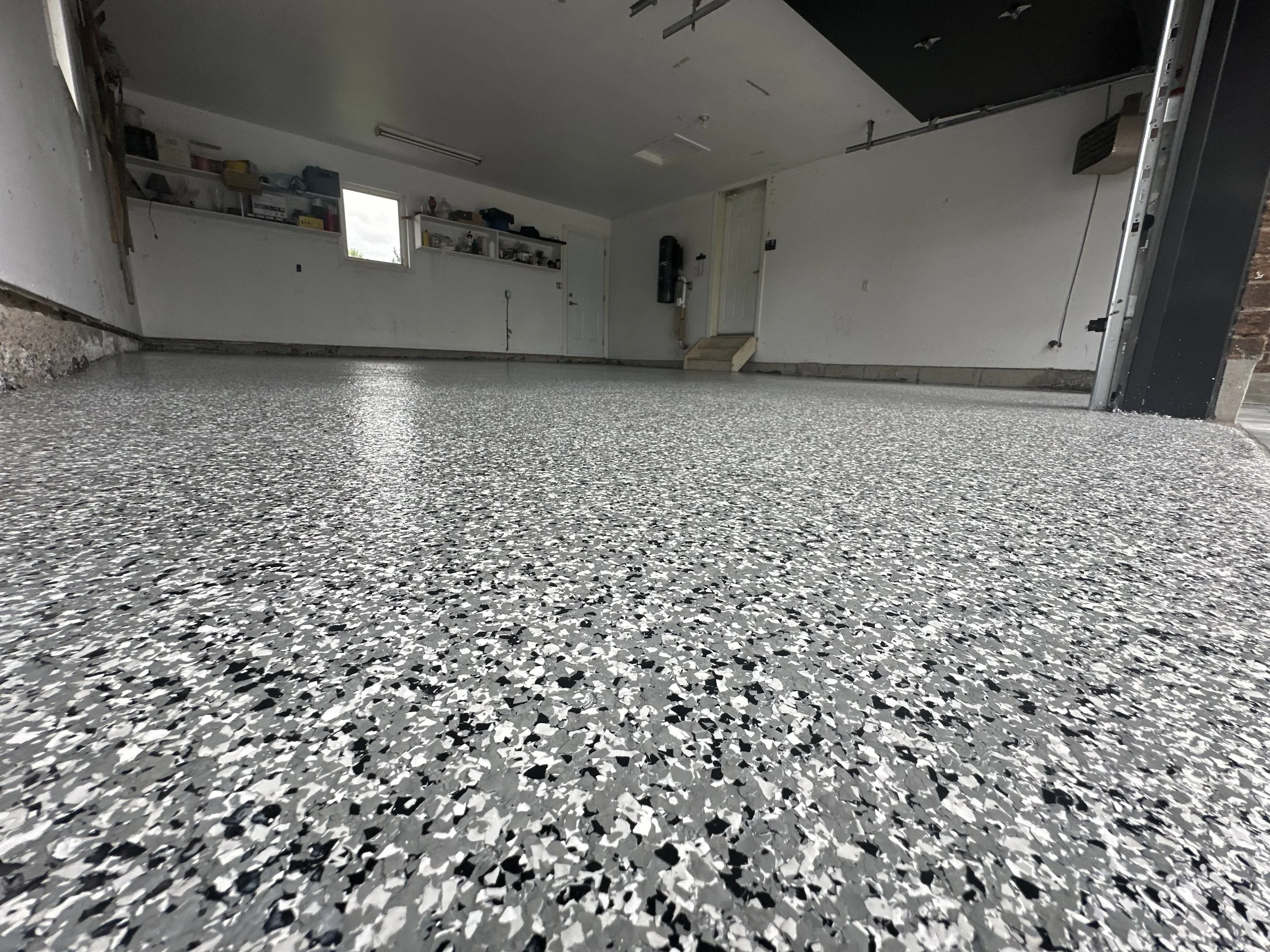 The image shows a clean, textured gray and white speckled epoxy garage floor with a white wall, shelves with storage containers, a window, and a door in the background.