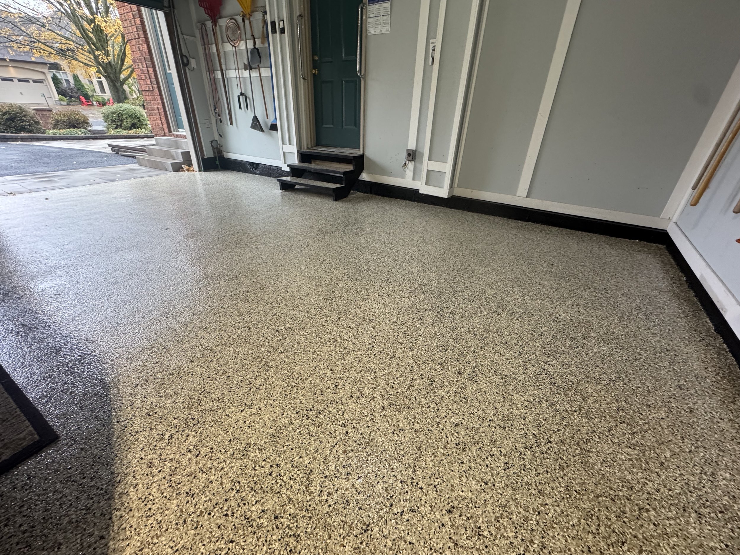 Clean garage with speckled epoxy floor, work tools hanging on wall, steps leading to a green door, and an open garage door showing a driveway outside.