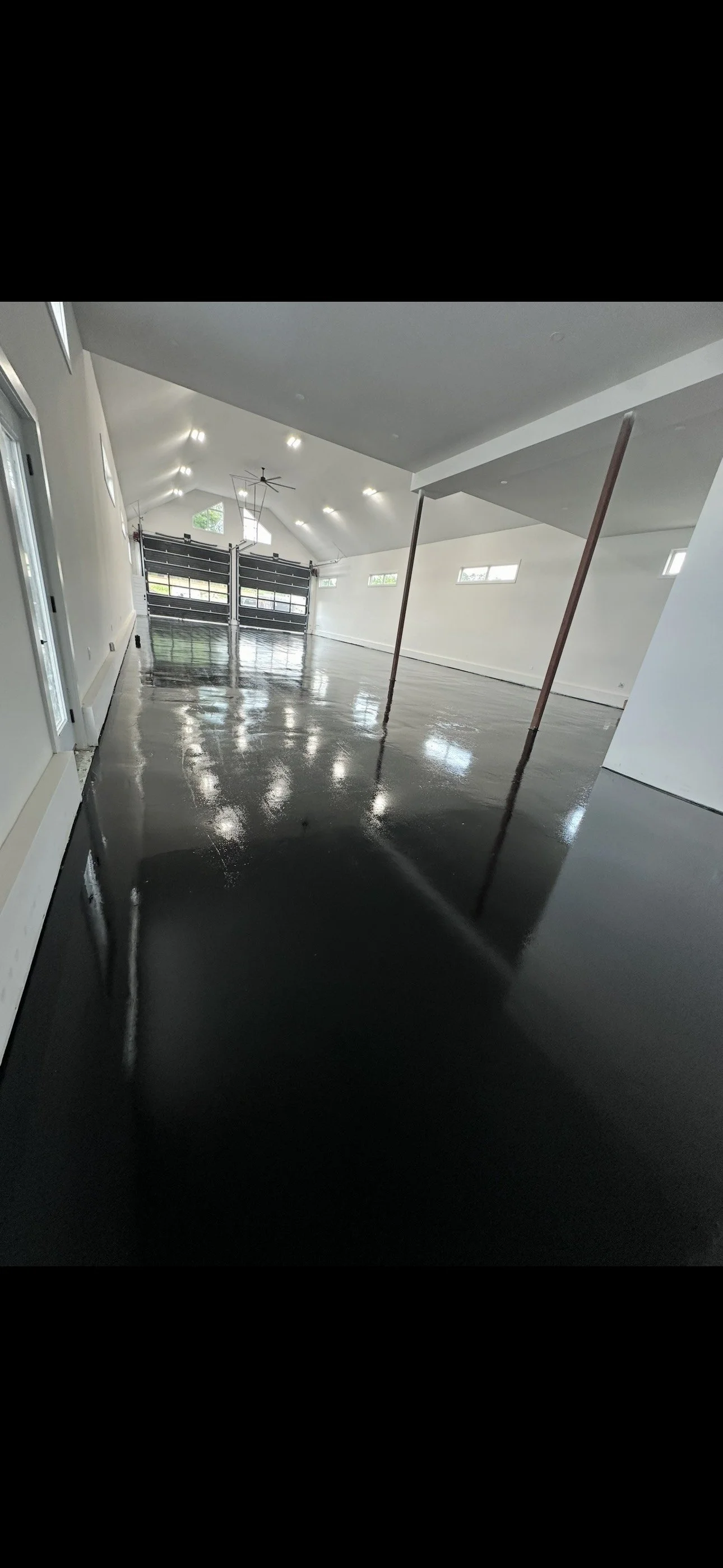 Interior view of a spacious, modern garage or multipurpose room with a black glossy epoxy floor, white walls, and a high ceiling with multiple skylights and a ceiling fan. Garage doors are visible at the far end.