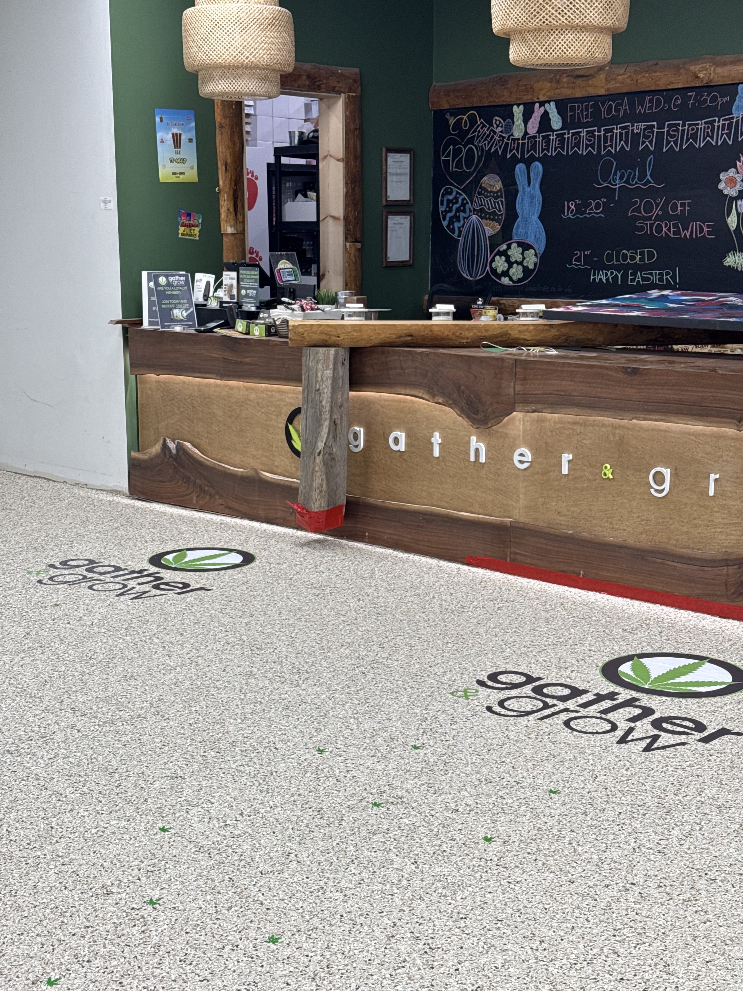Reception area of a store or cafe with a wooden counter, social distanced stickers on the floor with the logo and name 'gather & grow', a chalkboard with Easter-themed drawings, and a doorway leading to the kitchen.