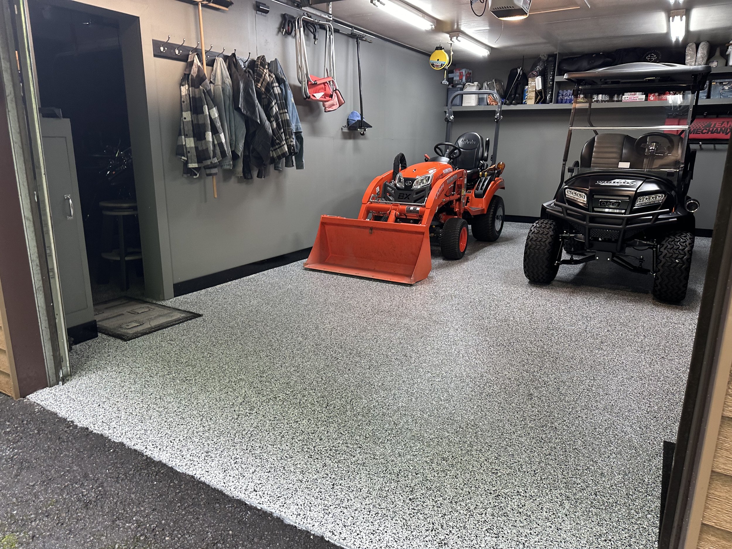 A garage with hanging jackets and bags on the wall, a small orange tractor with a front loader attachment, and a black utility vehicle parked inside.