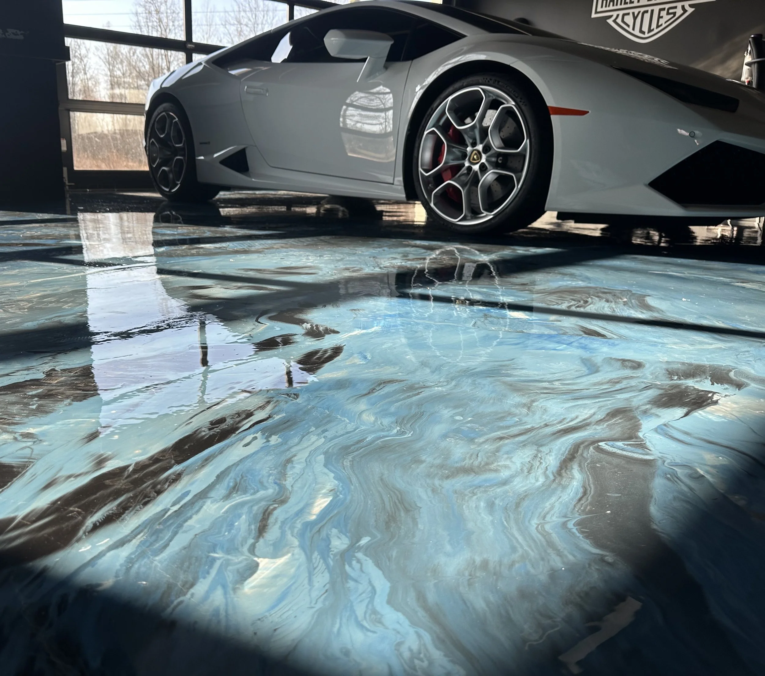 A silver Lamborghini sports car parked inside near a large window, with a black wall featuring a Harley-Davidson logo in the background. The floor is covered with a blue and gray marbled vinyl sheet.