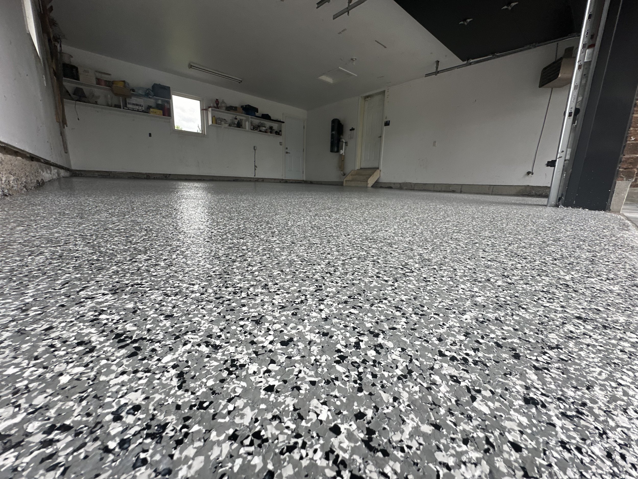 View of a garage floor with an epoxy coating, featuring a speckled gray, black, and white pattern, with garage walls, shelves, and a door in the background.