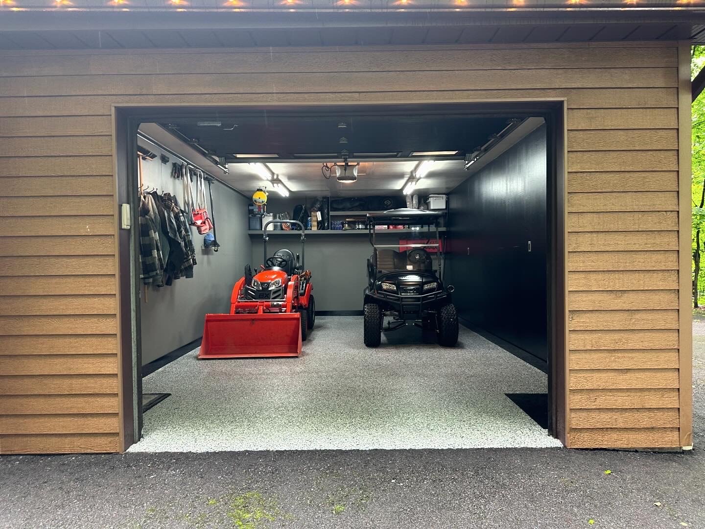 The image shows a garage with two small vehicles, a red tractor and a black utility vehicle, parked inside. The garage has wooden siding outside and a textured floor. There are hooks with clothing and hats on the left wall, and shelves with various i