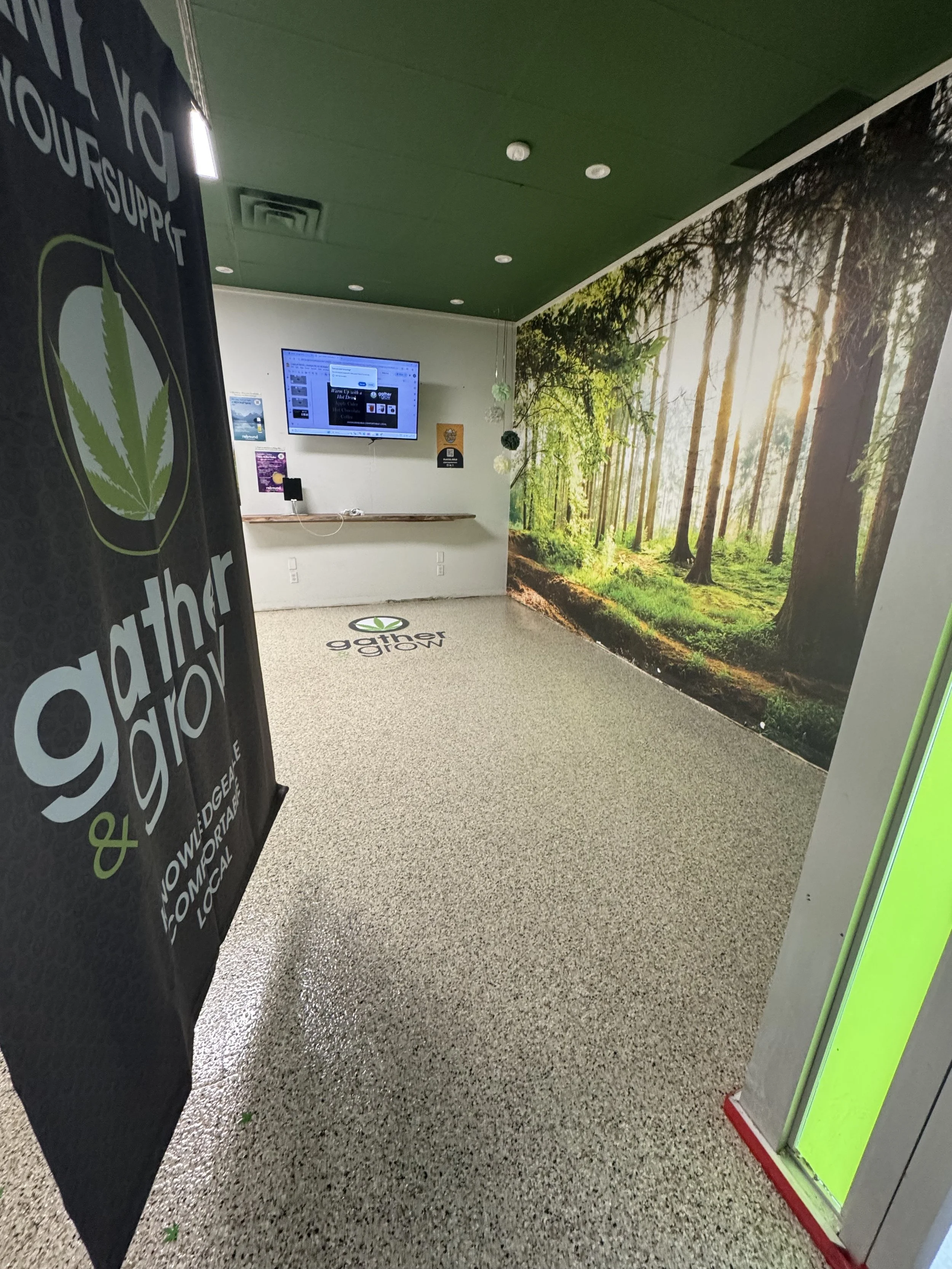 Interior of a cannabis dispensary with a forest mural on the wall, a TV screen, and a cart with branding for gather grow, focusing on cannabis products and environmental themes.