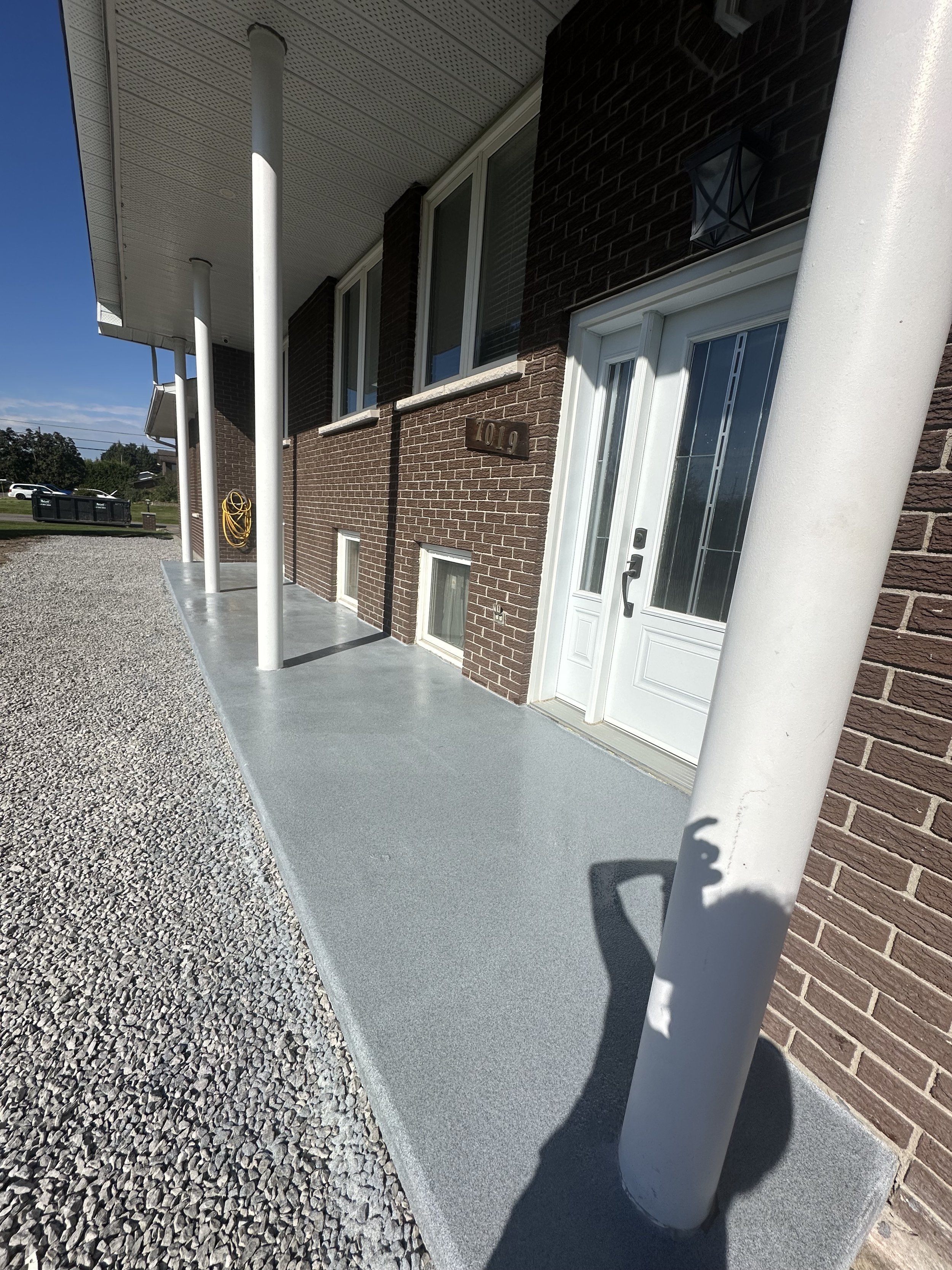 View of the front porch of a brick house with a concrete walkway, white support columns, white door, and exterior wall-mounted light fixtures.
