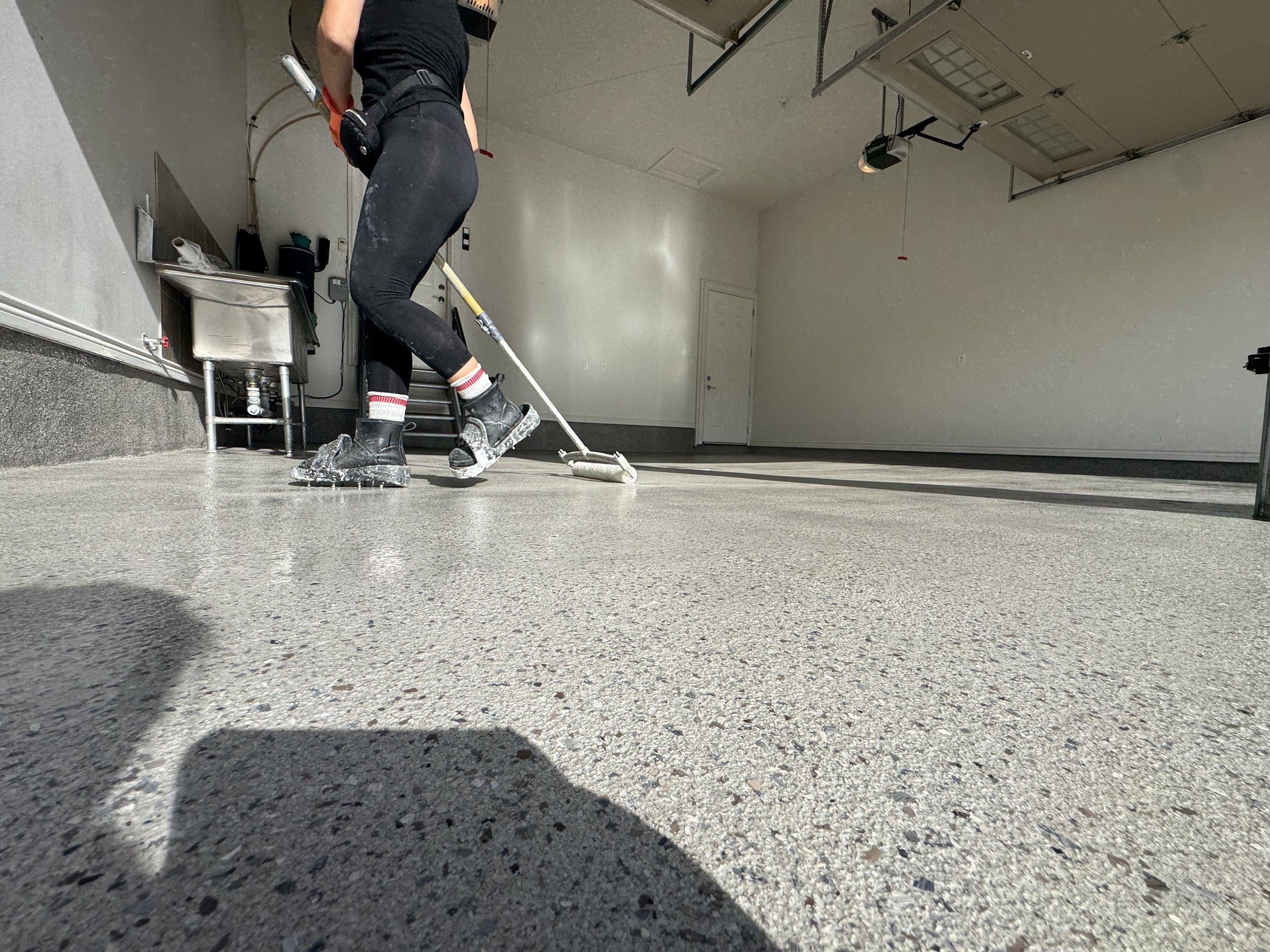 Person cleaning a garage with a floor mop, wearing black clothing, boots, and socks with red and white stripes, on a light gray textured concrete floor.