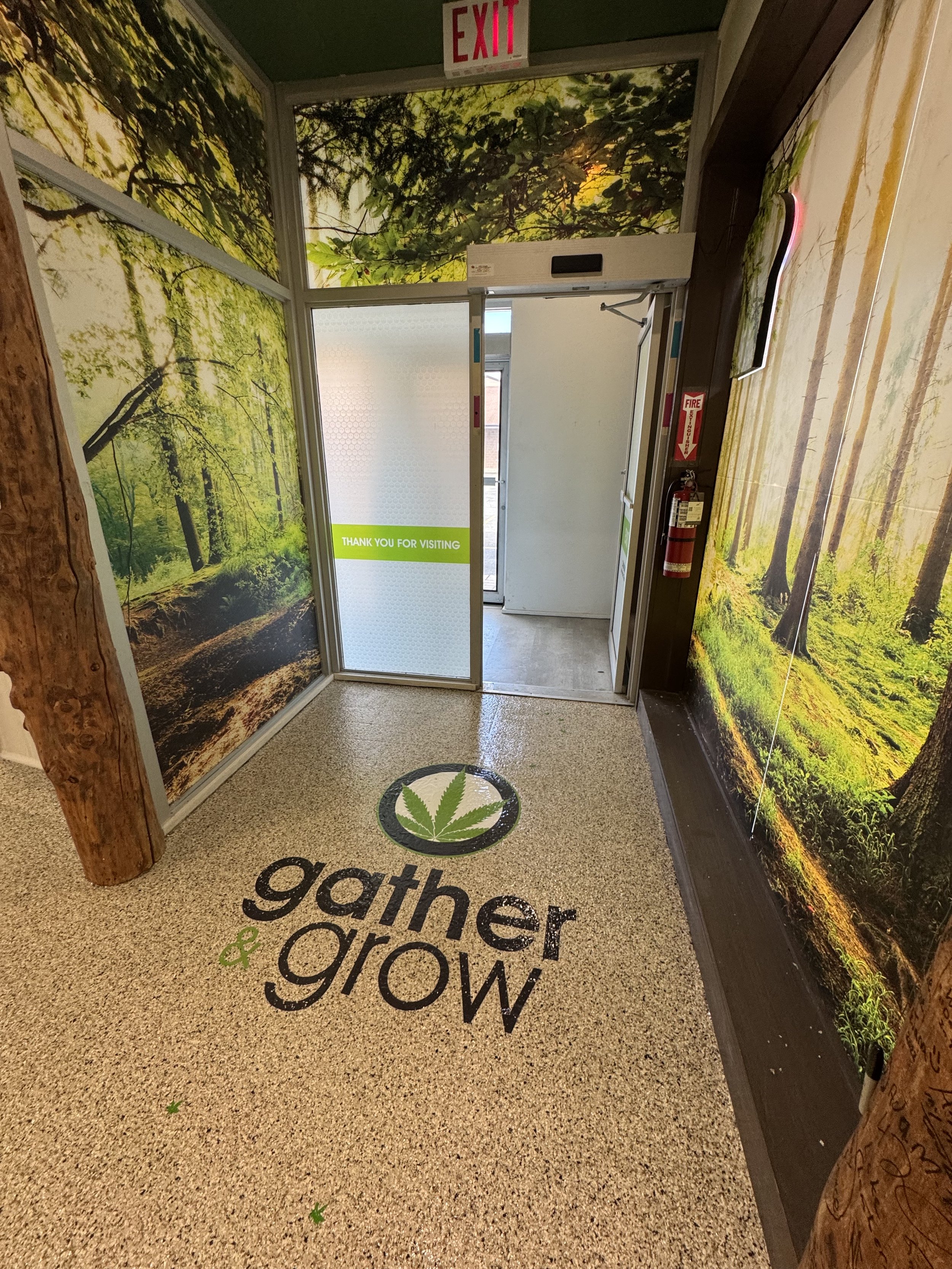 Interior of a building with walls covered in forest-themed murals showing trees and greenery. The floor has a logo with a cannabis leaf inside a circle and the words 'gather & grow'. There is an exit door with a sign that says 'Thank you for visiting