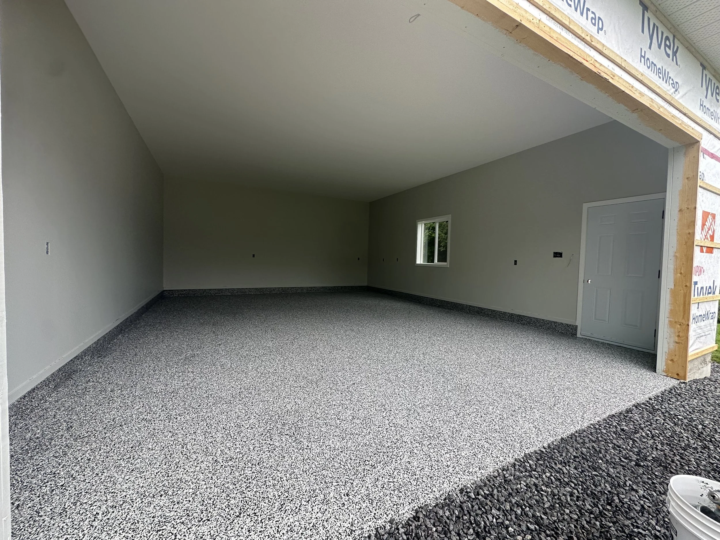 Inside a building under construction showing an unfinished garage with speckled gray flooring, white walls, a small window, and an open doorway.