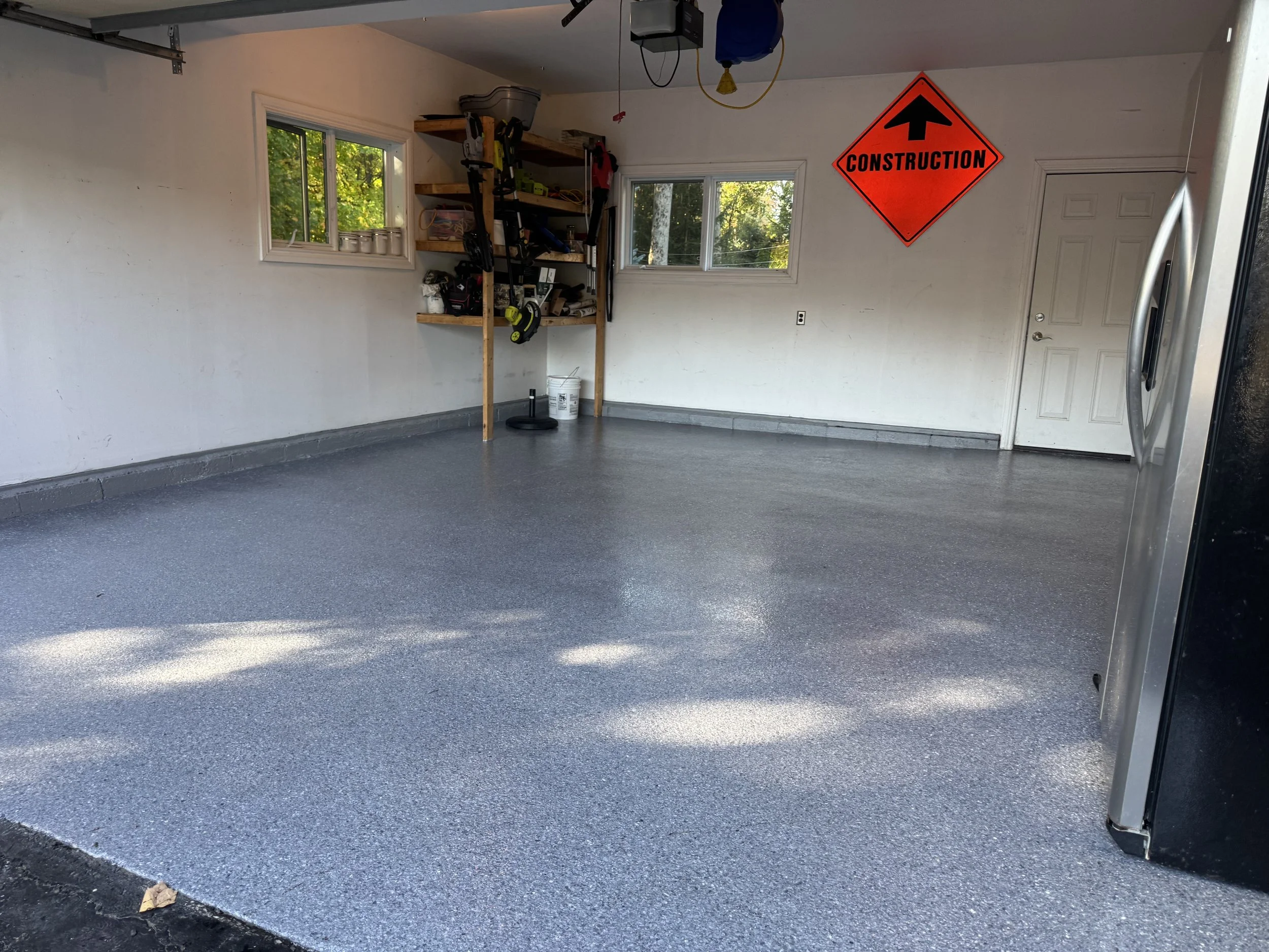 Empty garage with gray epoxy floor, white walls, two small windows, a door, a workbench with tools, and a bright orange construction sign on the wall.