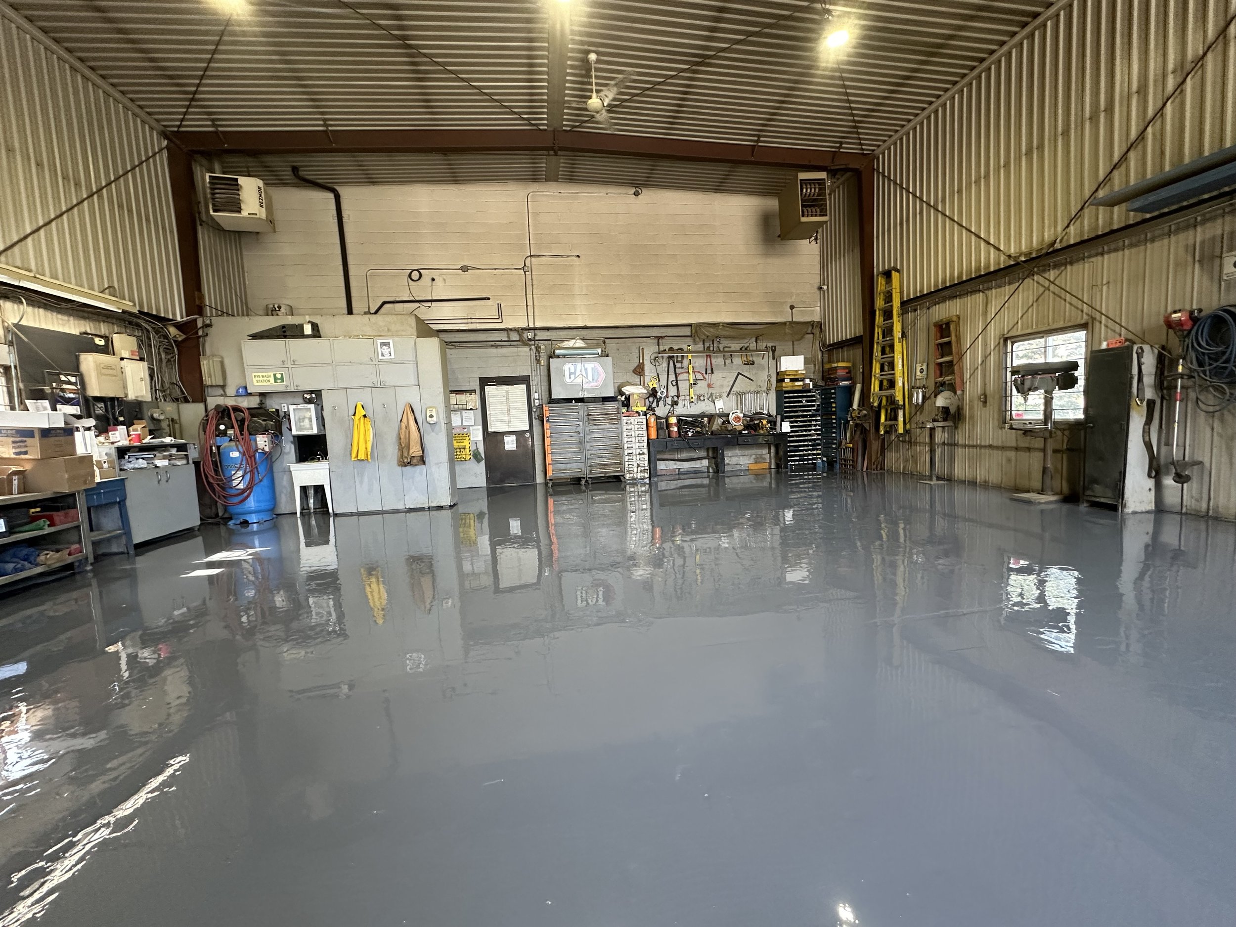 Inside a garage or workshop with newly applied gray epoxy flooring, various tools, equipment, and storage units are visible against the walls, with two small windows providing natural light.