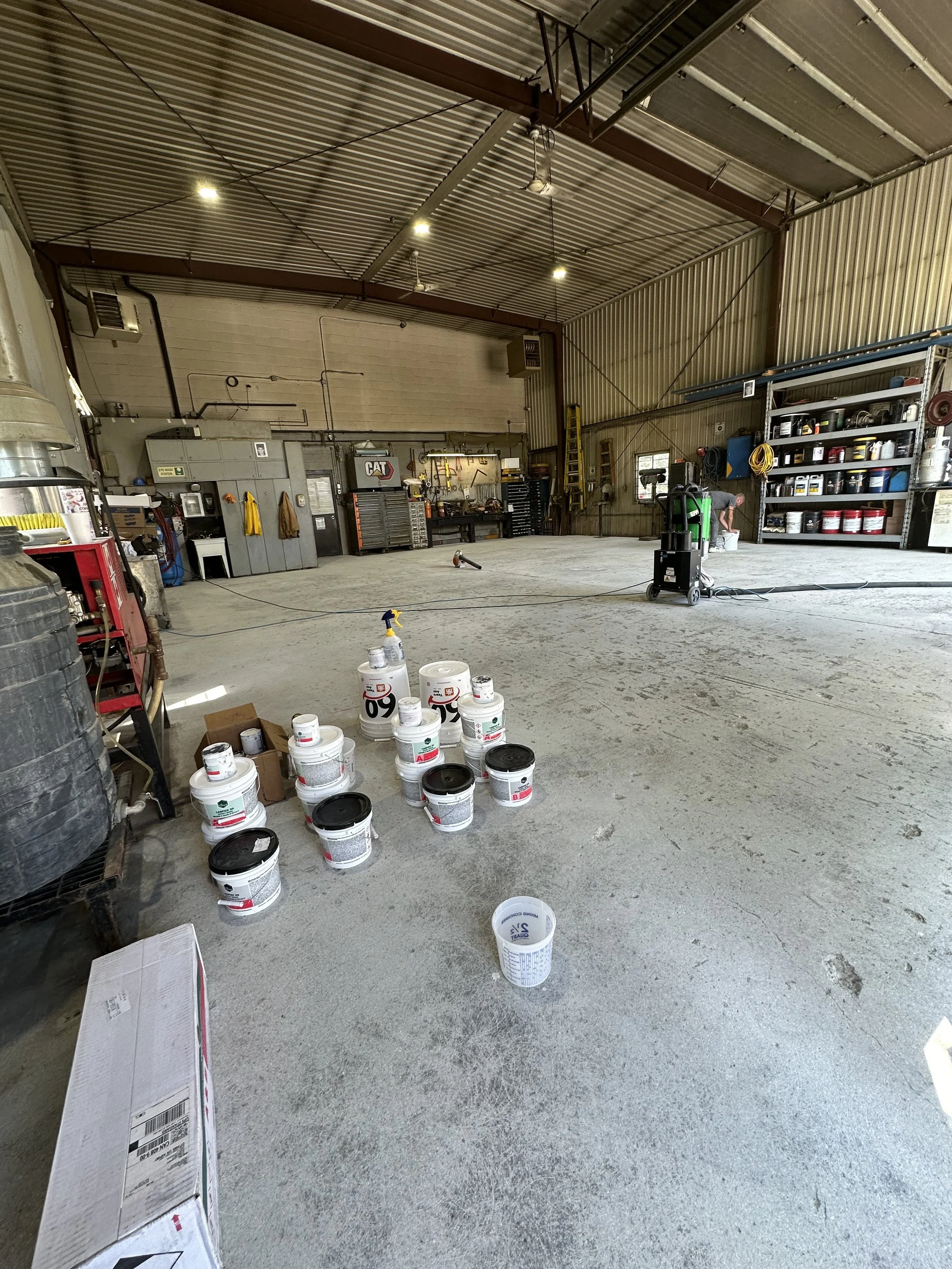 Interior of a large workshop or garage with various tools, shelves, and equipment. Several buckets and containers are on the floor in the foreground, and a person working near the shelves on the right side.