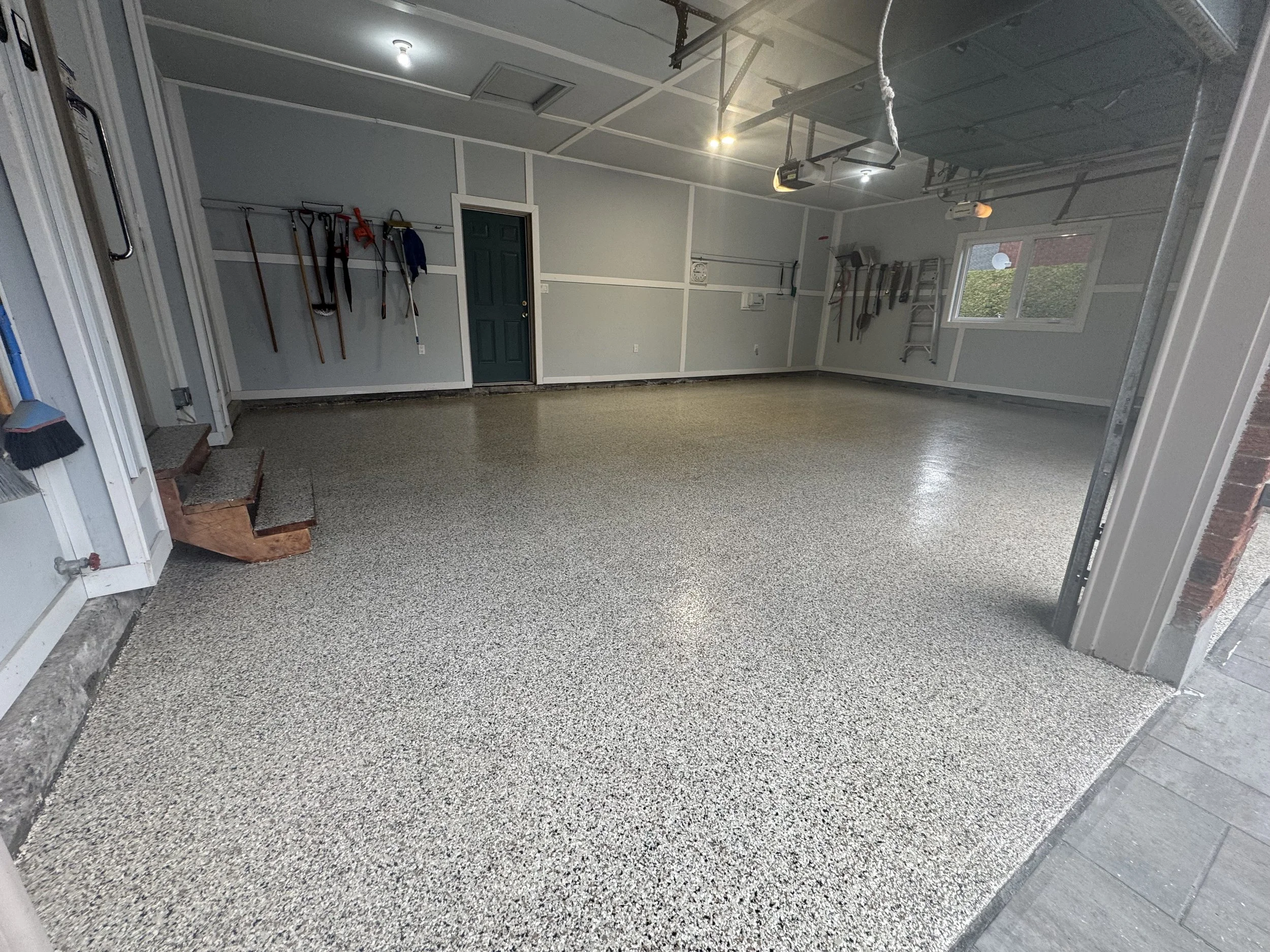 An empty garage with epoxy-coated floor, hanging tools on the walls, a small set of stairs, and windows providing natural light.