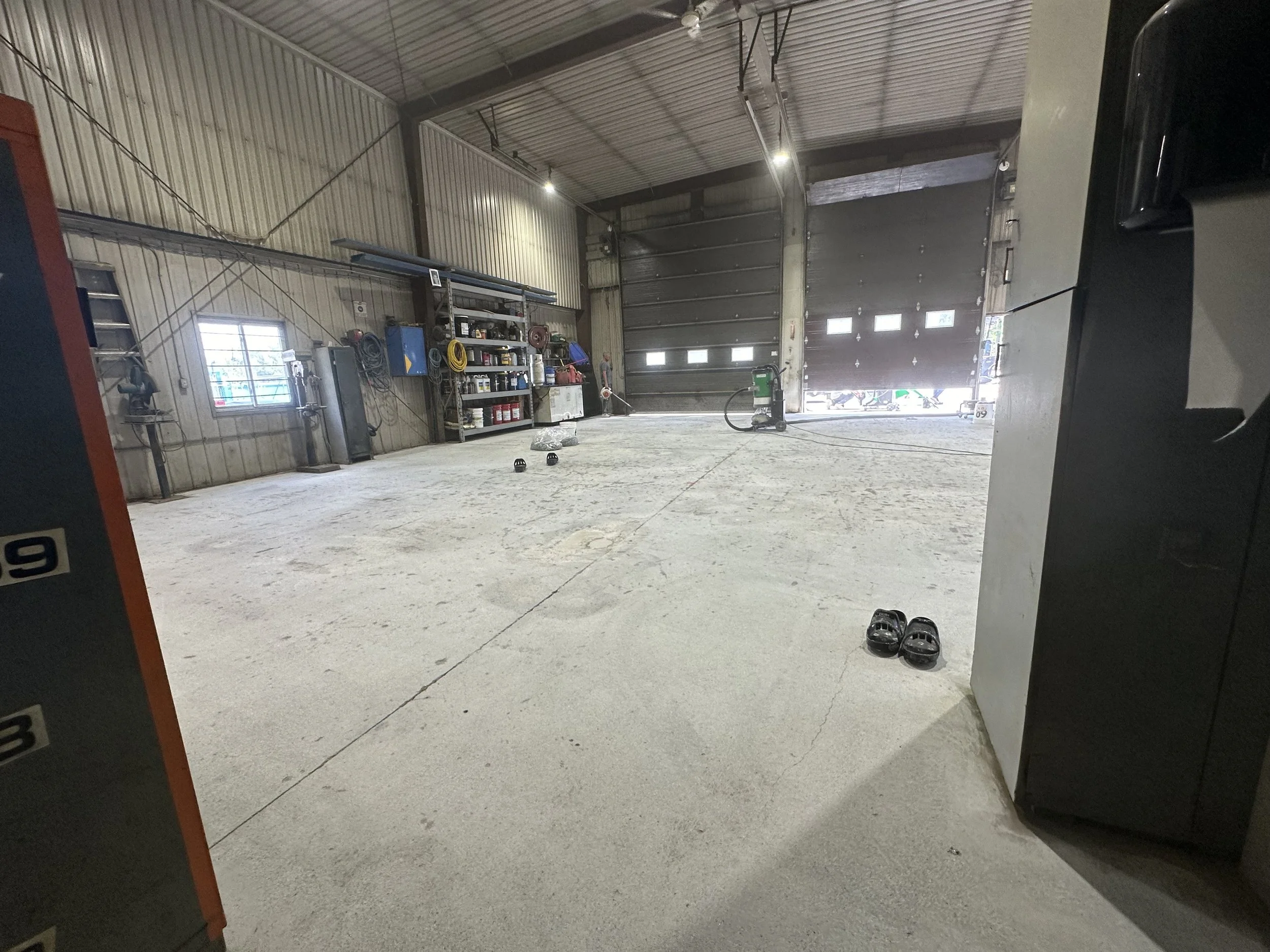 Interior of a spacious garage or workshop with a concrete floor, metal walls, and a large roll-up door at the back. Shelves with tools, supplies, and equipment are on the left side, and there are scattered black shoes and a pair of shoes on the right