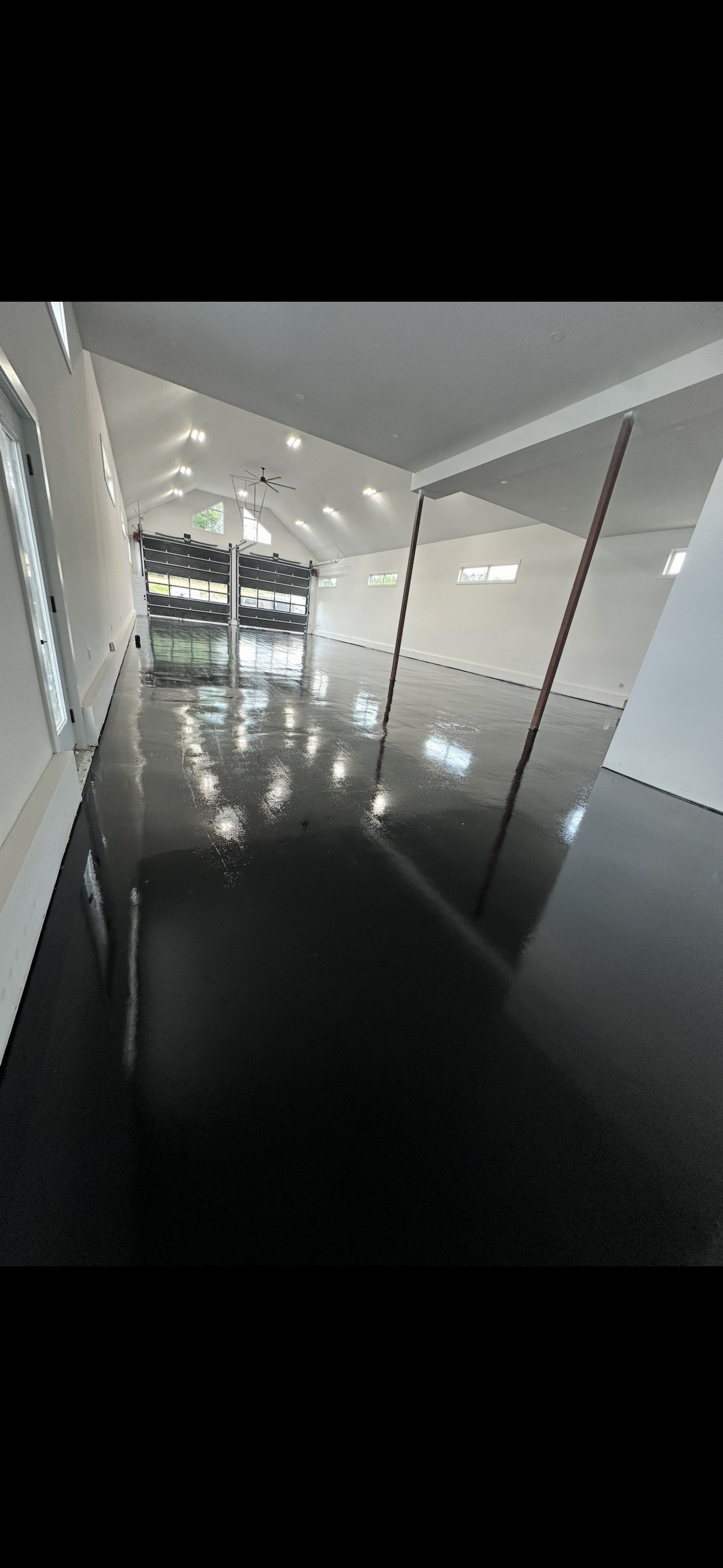 An empty, newly finished garage with a polished black concrete floor reflecting light. The walls are white, and there are several small windows near the ceiling. The garage has a high, vaulted ceiling with a ceiling fan and recessed lighting. Two gar