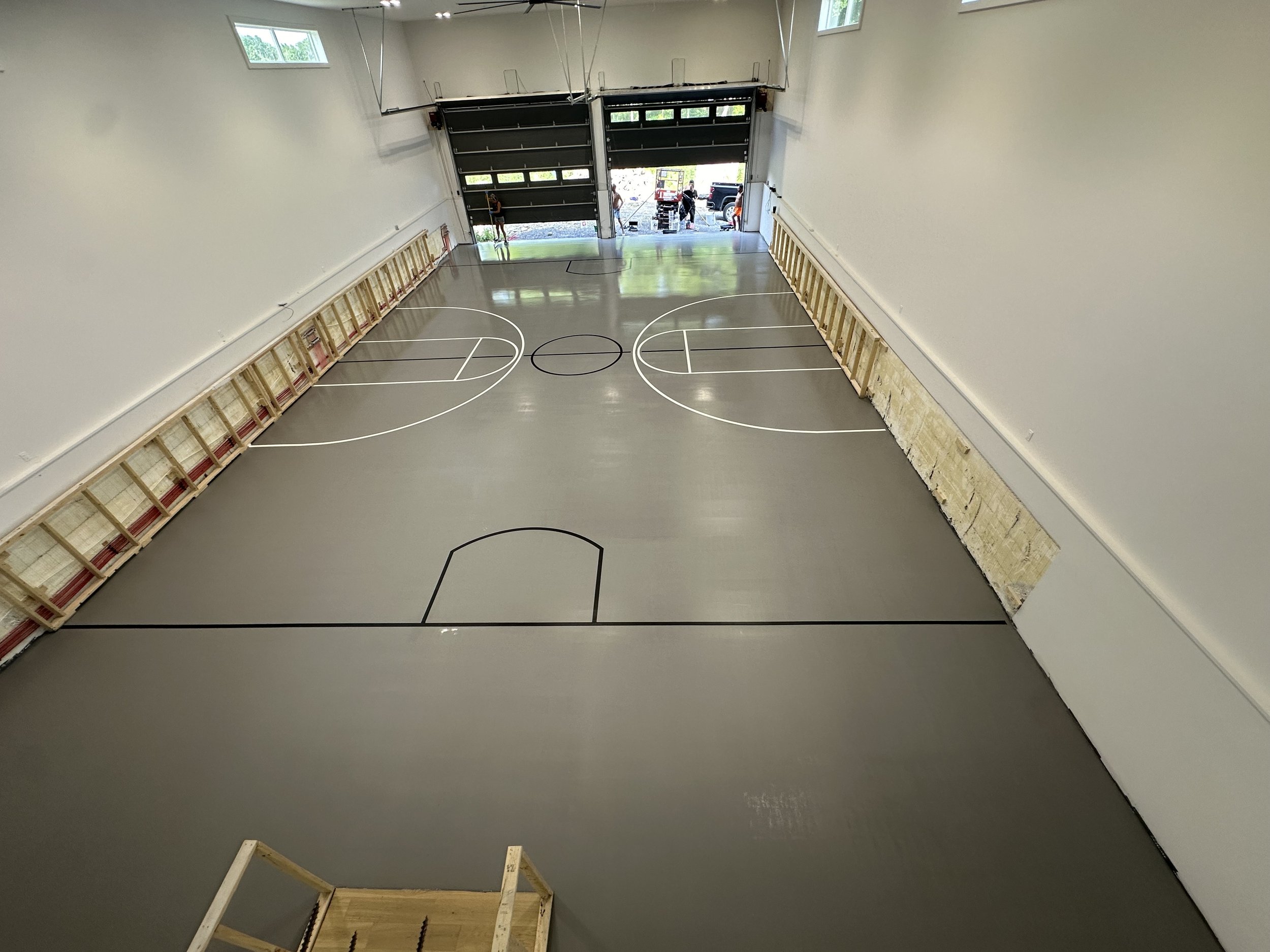 An indoor basketball court under construction with a polished gray floor, black and white court lines, wooden benches along the side walls, and a large open garage-style door at the far end with people outside.
